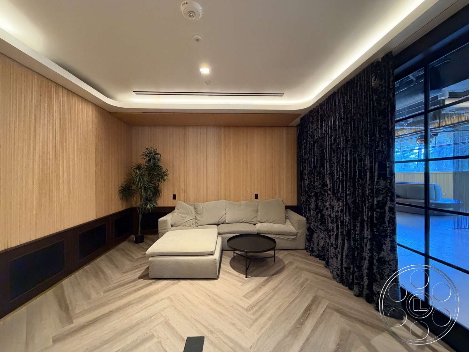 Office 130 - modern coffee table, herringbone pattern flooring, black framed glass doors, recessed ceiling lighting, upholstered sectional sofa, indoor plant accents, wood paneling walls, fabric drapery