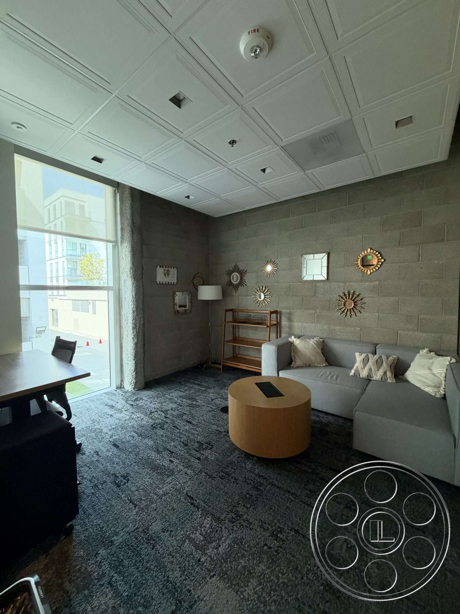 Office 130 - neutral color palette, large windows, concrete block walls, gray sectional sofa, round wooden coffee table, modern floor lamp, open floor plan layout, textured carpet flooring, industrial-style decor, decorative wall mirrors, dropped ceiling tiles
