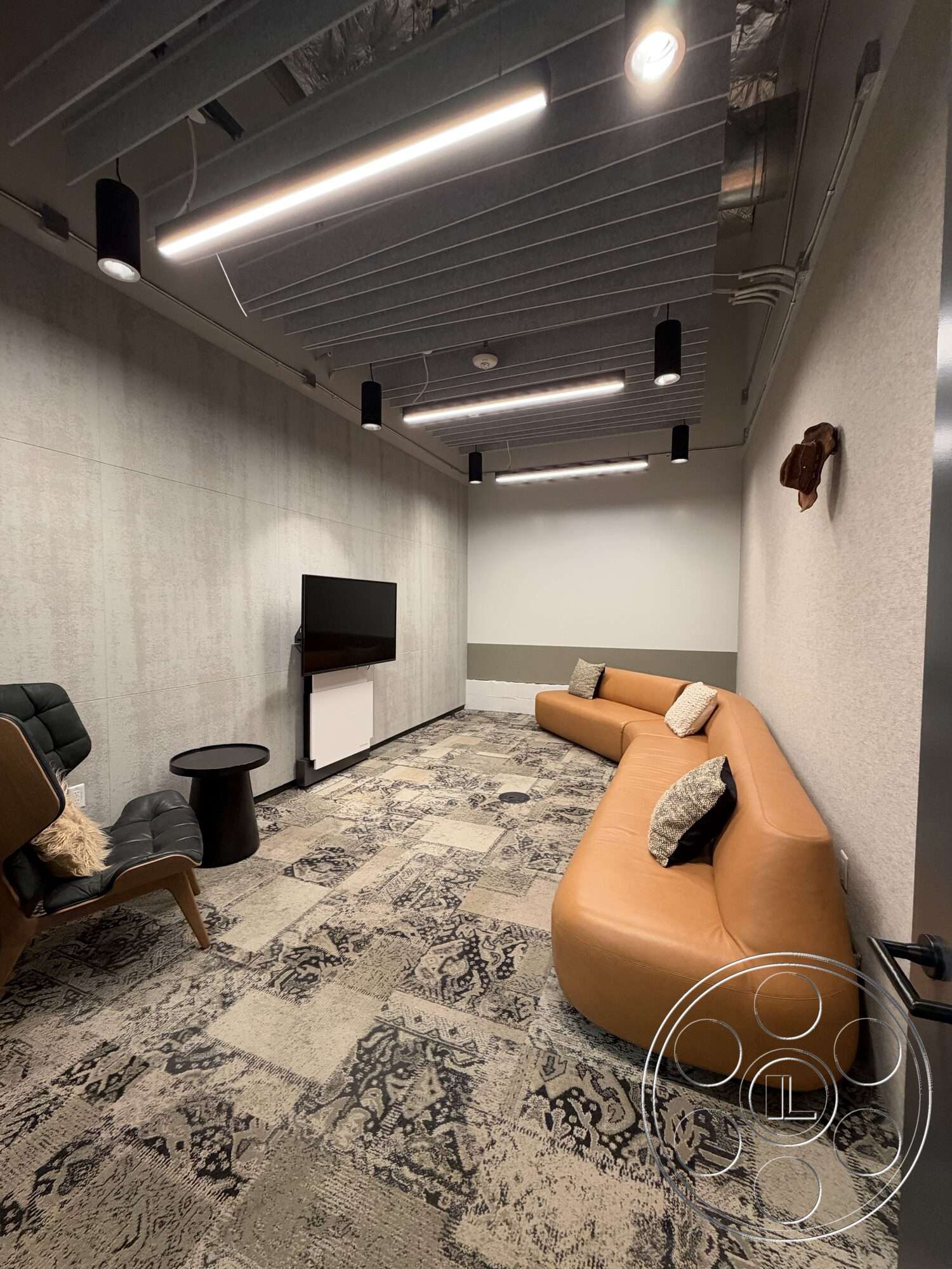 Office 130 - carpet flooring, decorative wall art, textured plaster walls, recessed ceiling lights, wood accent table, modern wall-mounted TV, curved leather sofa, exposed ceiling ducts, black lounge chair