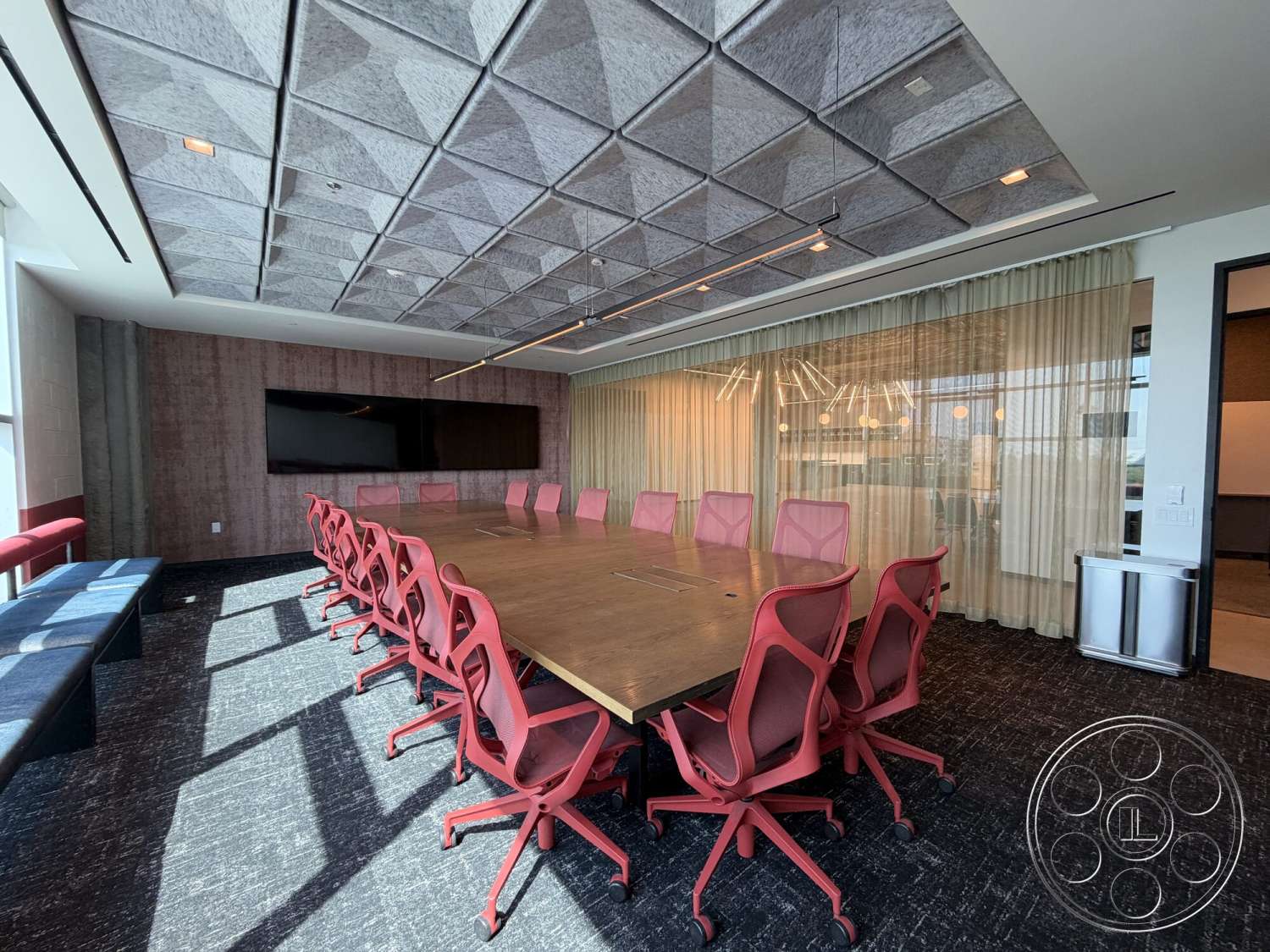 Office 130 - wall-mounted television, modern pendant lighting, carpeted flooring, curtained windows, glass partition walls, acoustic ceiling panels, wood conference table, textured fabric ceiling tiles, red ergonomic office chairs, open layout meeting space