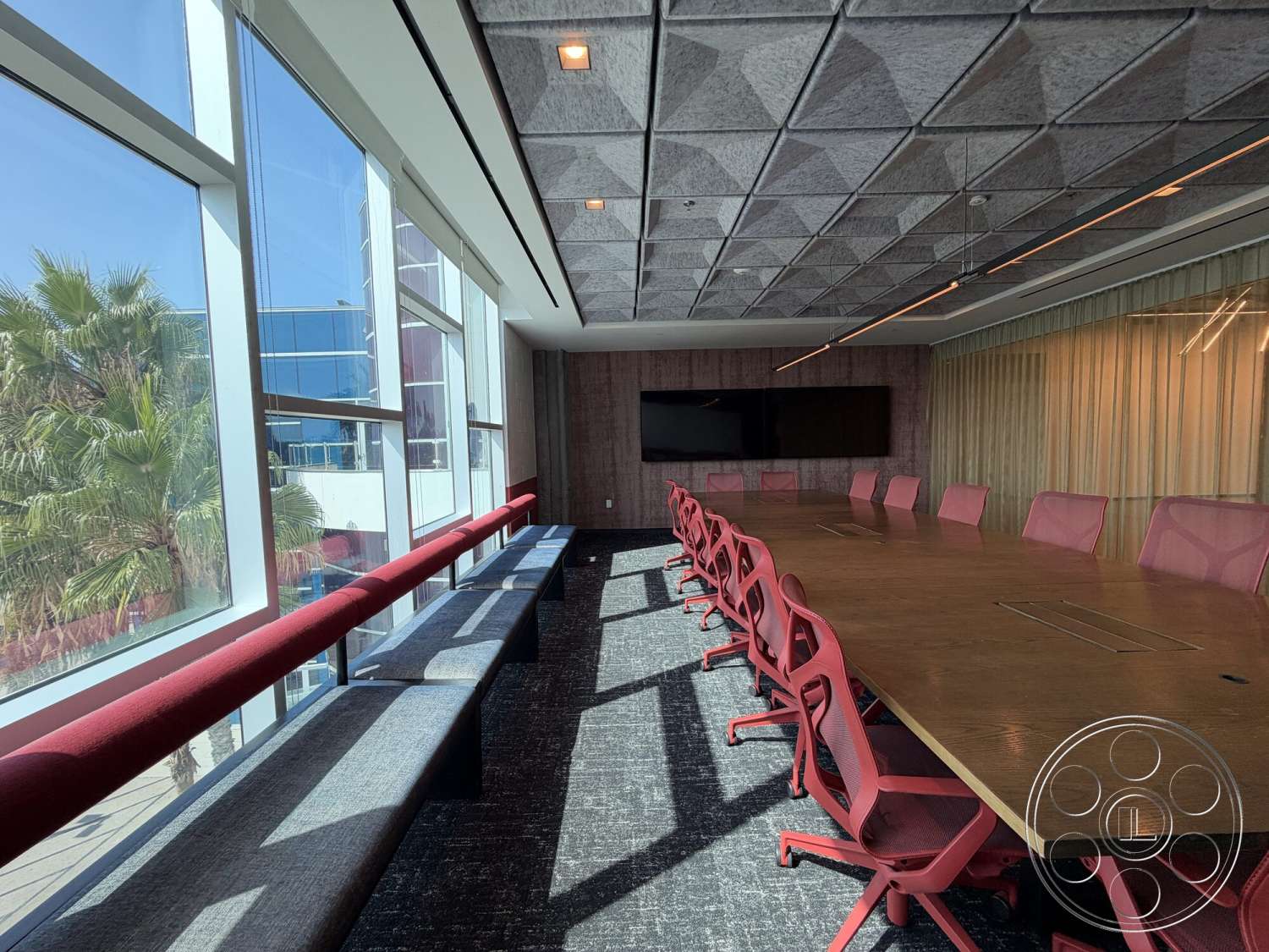 Office 130 - glass windows, textured wall paneling, red upholstered chairs, acoustic ceiling tiles, flat-screen wall-mounted television, fluorescent ceiling lights, wood conference table, fabric bench seating, gray carpet tiles