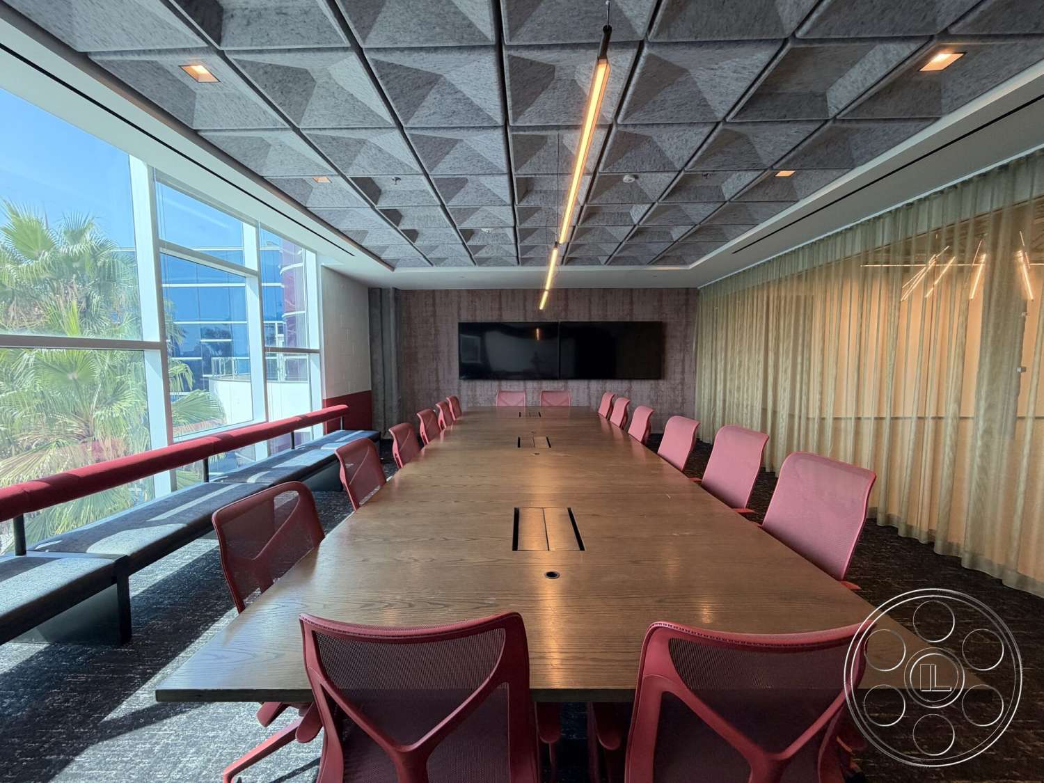 Office 130 - large glass windows, wood accent wall, flat-screen wall-mounted television, textured ceiling panels, dark carpet flooring, wood conference table, pink upholstered conference chairs, vertical window blinds, soft LED lighting, custom built-in bench seating
