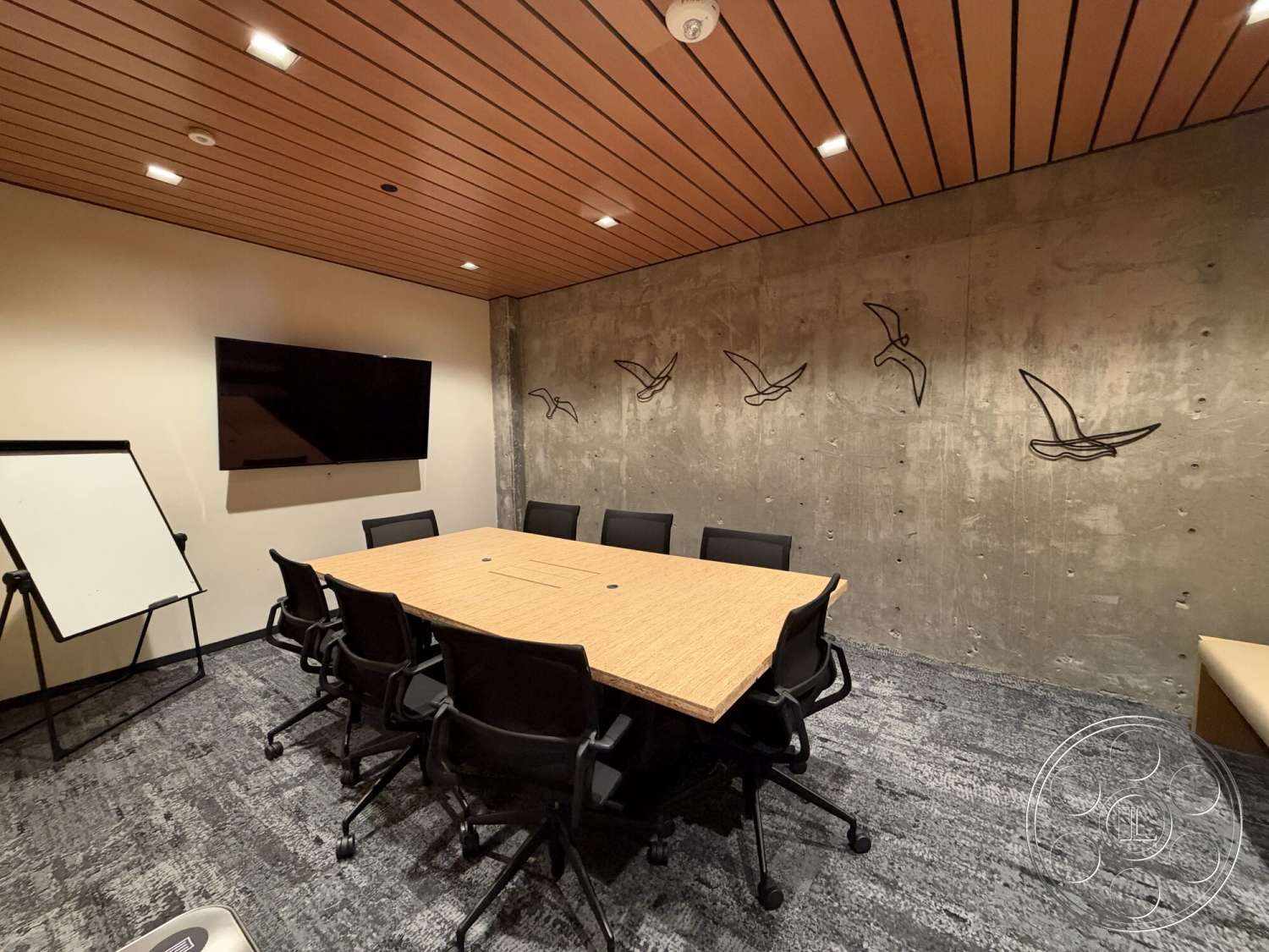 Office 130 - concrete wall, wood ceiling panels, black office chairs, textured carpet flooring, ambient recessed lighting, rectangular conference table, wall-mounted flat screen TV, metal wall art, foldable whiteboard