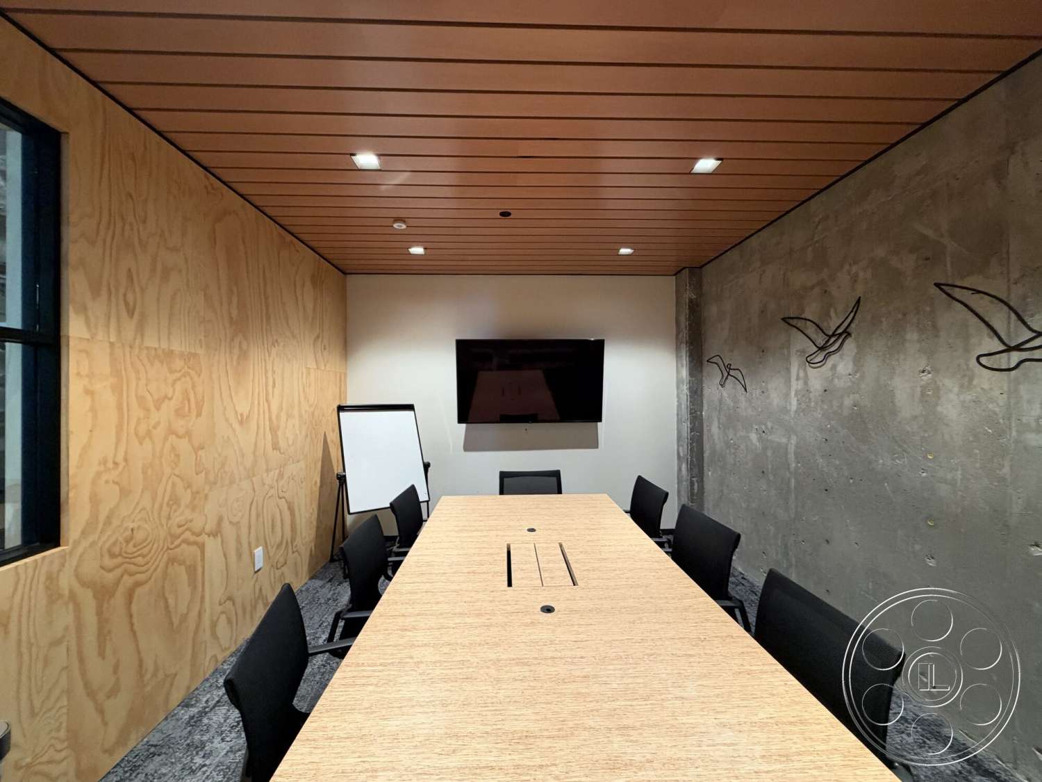 Office 130 - exposed concrete wall, glass window, gray carpet flooring, black office chairs, rectangular conference table, slatted wood ceiling, plywood wall paneling, built-in television mount