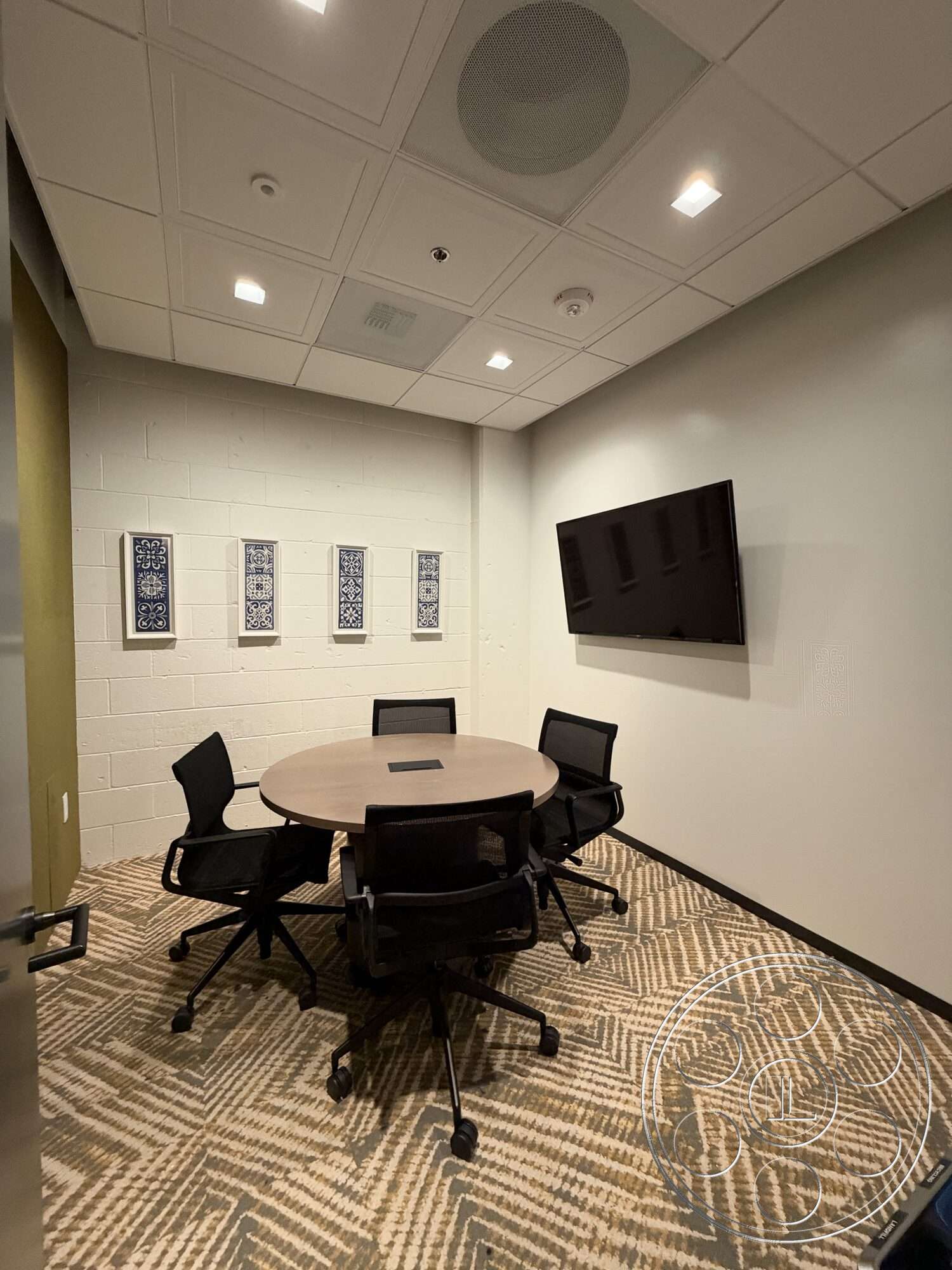 Office 130 - acoustic ceiling tiles, recessed lighting fixtures, exposed concrete accent wall, black ergonomic office chairs, carpet flooring in herringbone pattern, drywall walls with white paint, built-in television wall, wooden round conference table, decorative wall art with geometric patterns, ceiling-mounted speaker system