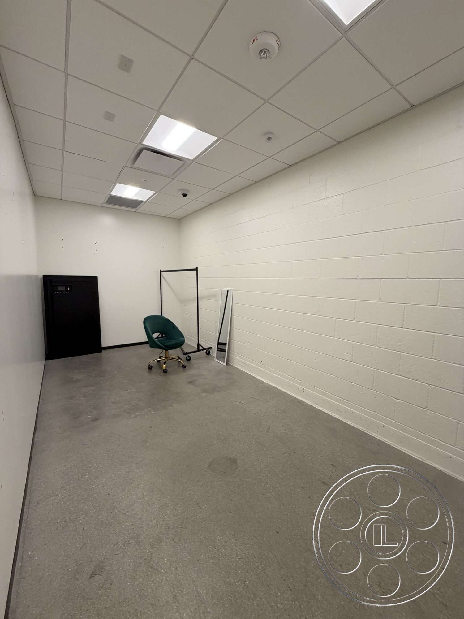 Office 130 - minimalist design, concrete floor, commercial space, empty room, full-length mirror, white painted walls, drop ceiling with fluorescent lighting, black storage cabinet, green rolling office chair