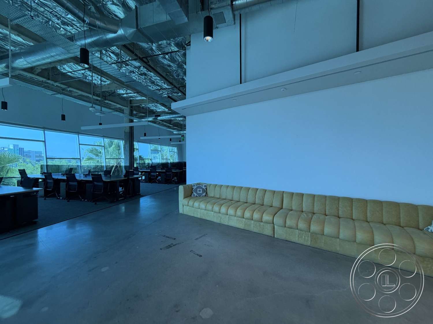 Office 130 - floor-to-ceiling windows, exposed concrete flooring, industrial ceiling with exposed ductwork, minimalist white walls, curved yellow modular sofa, modern office workspace, black office desks, large open office layout