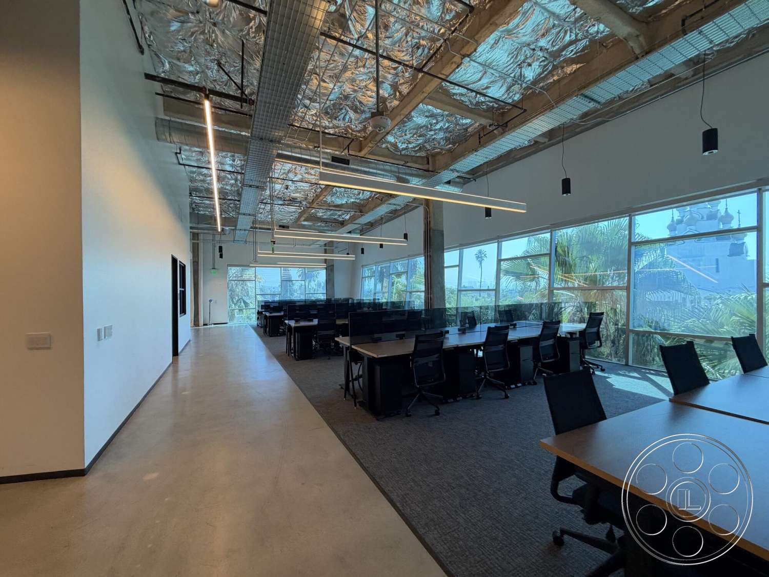 Office 130 - polished concrete flooring, minimalist design elements, modern pendant lighting, floor-to-ceiling windows, palm tree view, open office layout, glass partition walls, industrial-style architecture, textured carpet tiles, black ergonomic office chairs, exposed concrete ceiling beams, dark wood conference tables