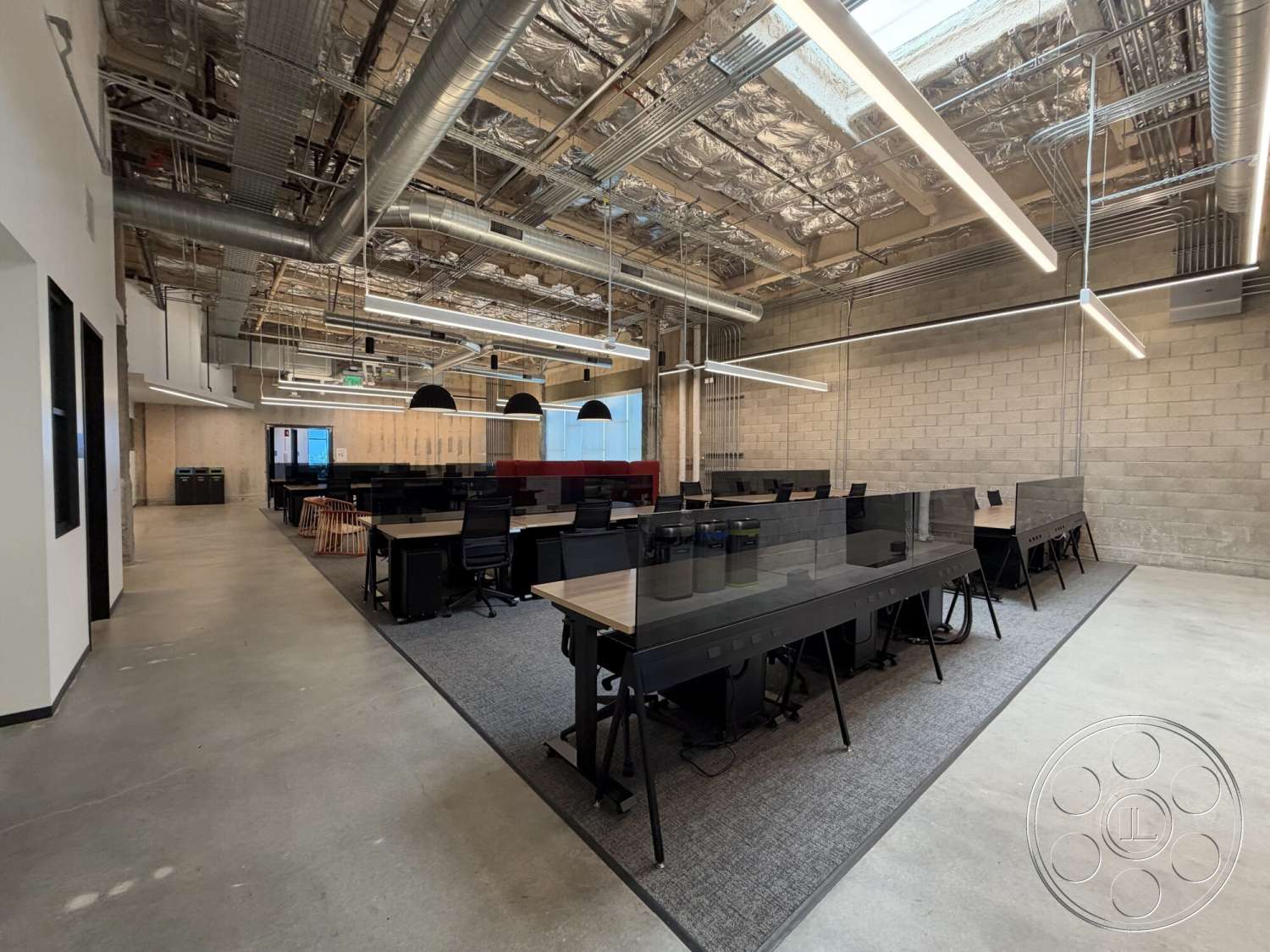 Office 130 - concrete flooring, large windows for natural light, exposed brick walls, open workspace, black office chairs, modern office environment, exposed ductwork ceiling, rectangular office layout, industrial-style lighting, task lighting fixtures, gray carpet tiles, glass desk dividers, plywood accent walls, red fabric seating area, high ceiling with metal beams