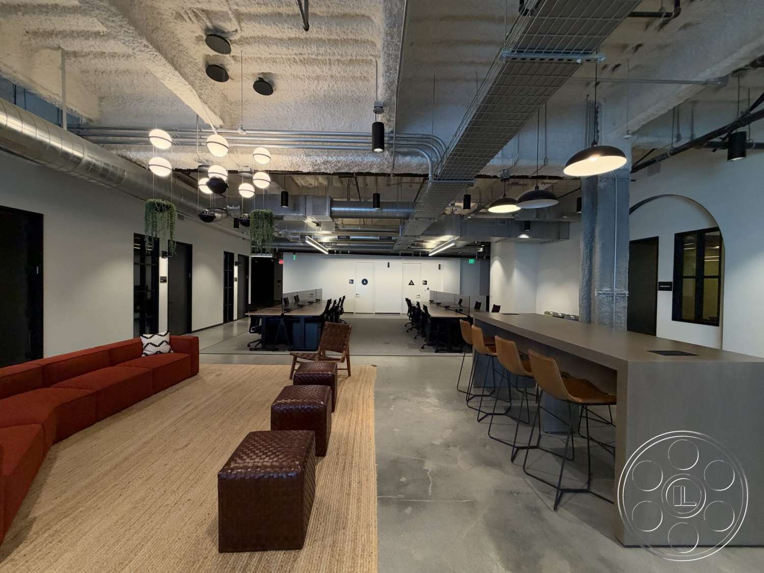 Office 130 - concrete flooring, modern pendant lighting, woven area rug, exposed concrete ceiling, metal ductwork, glass partition walls, industrial style architecture, open workspace layout, dark wood bar counter, upholstered red sectional sofa, wooden accent stools