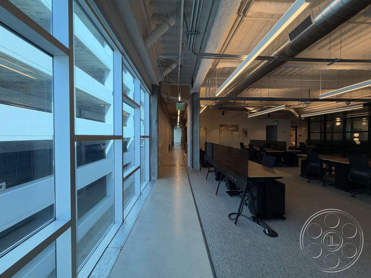 Office 130 - exposed concrete walls, open office layout, natural light from floor-to-ceiling windows, glass office partitions, light gray carpet flooring, metal ceiling beams, concrete hallway flooring, LED linear ceiling lights