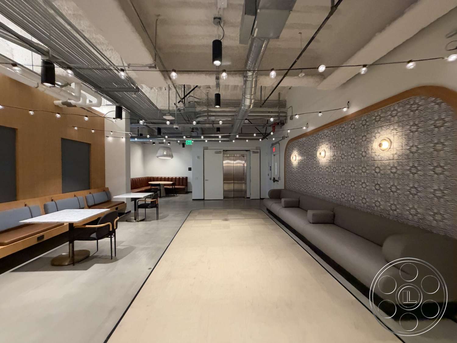 Office 130 - neutral color palette, open floor plan, concrete flooring, cozy seating area, minimalist design elements, industrial style, wood paneling accent wall, soft seating area, rectangular tables, exposed ceiling ductwork, round ceiling light fixtures, built-in booth seating, wall-mounted decorative lighting, modern lounge space