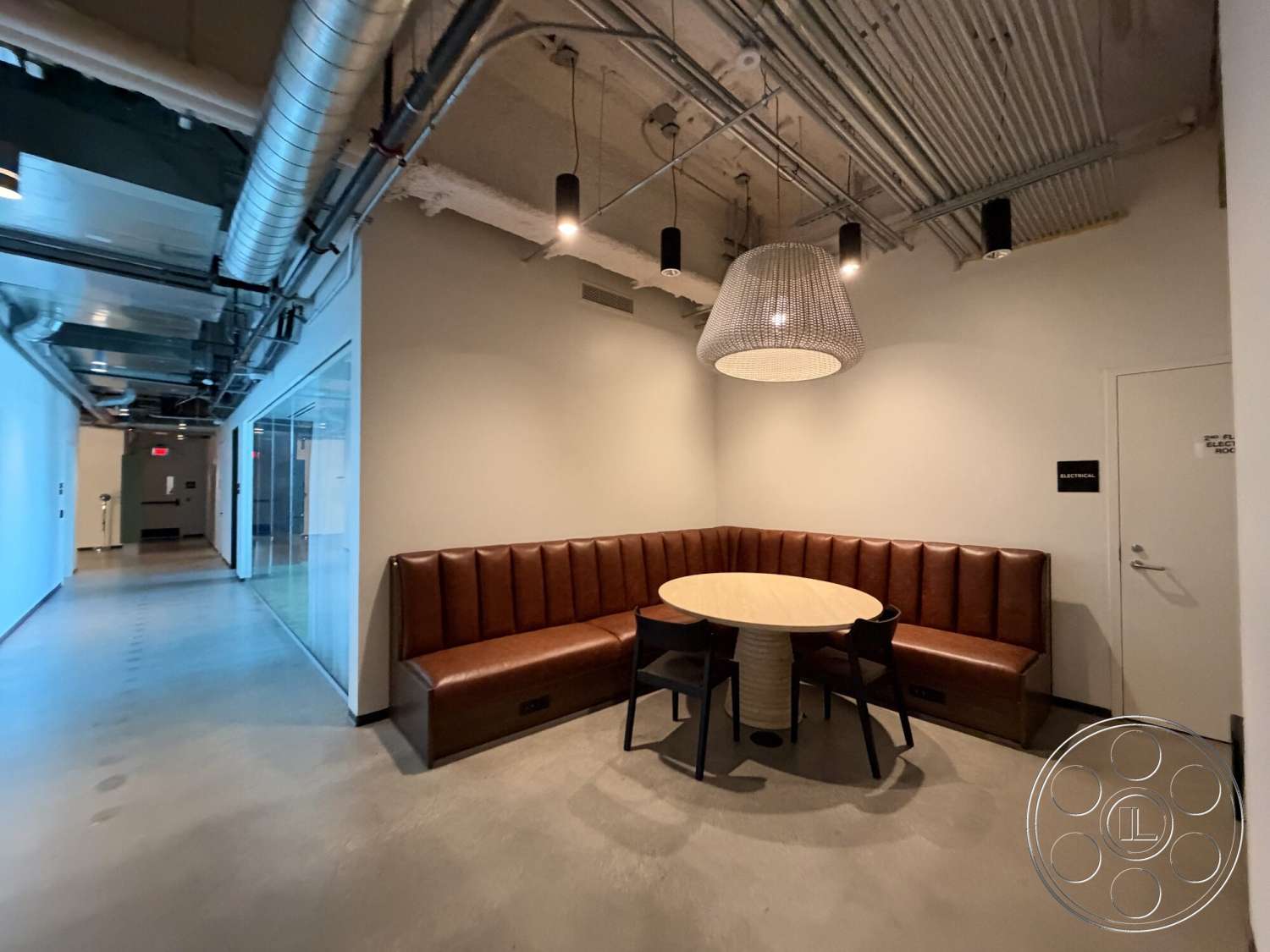 Office 130 - round table, polished concrete flooring, pendant light fixture, white painted walls, exposed ductwork ceiling, glass partition wall, built-in bench seating, modern office lounge area, brown leather upholstery