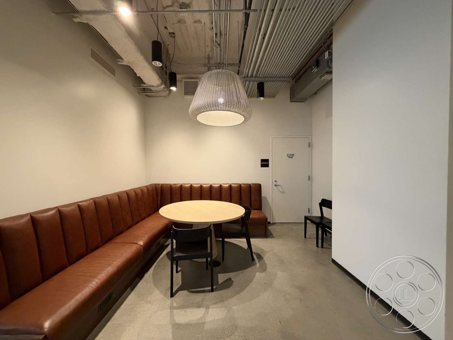 Office 130 - minimalist design, black metal chair, concrete flooring, soft ambient lighting, round wooden dining table, modern pendant light fixture, white painted walls, exposed ceiling with visible ductwork, leather upholstered booth seating