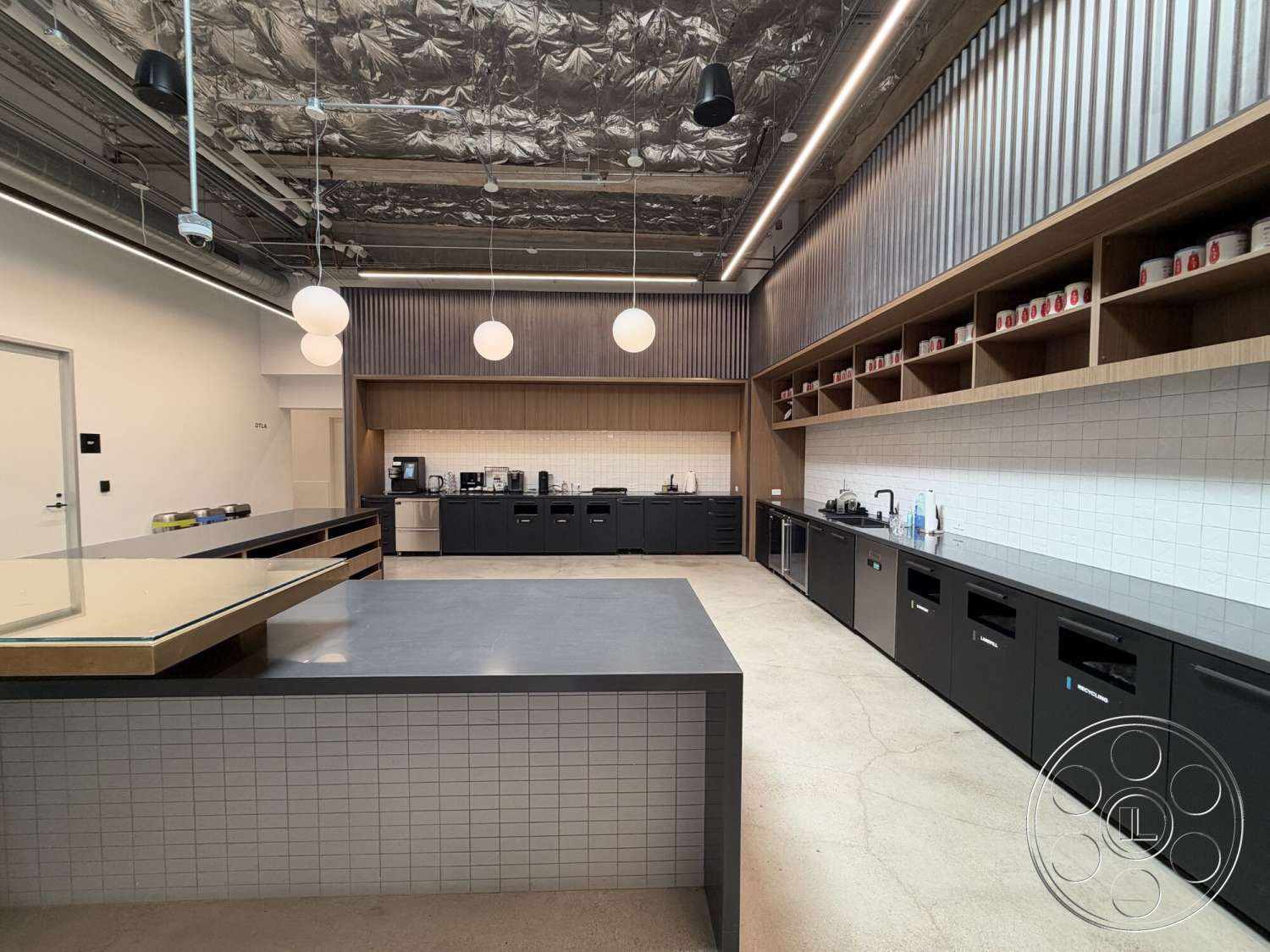 Office 130 - concrete flooring, black cabinetry, open kitchen layout, rectangular kitchen island, interior kitchen, wood accent shelves, metal ceiling tiles, white tile backsplash, industrial style kitchen, Hanging pendant lights
