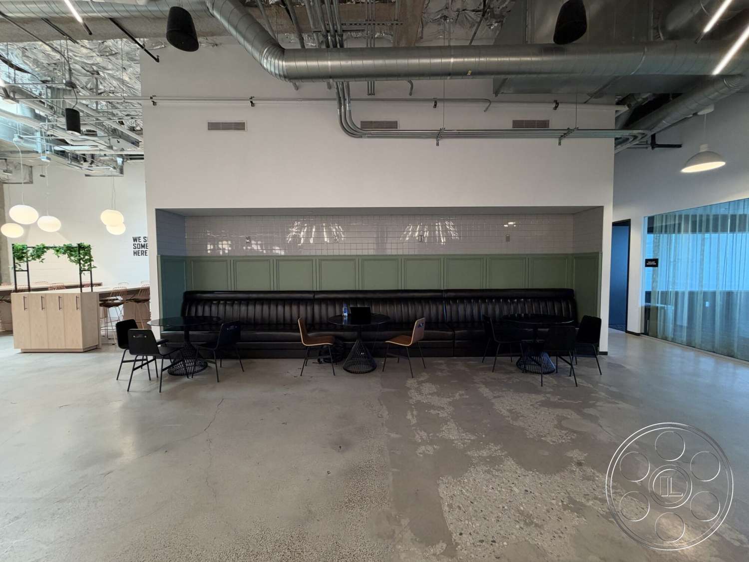 Office 130 - polished concrete flooring, black metal chairs, open office layout, minimalist design aesthetic, decorative plant accents, exposed ductwork ceiling, industrial-style lighting fixtures, black leather upholstered bench, green paneling wall, white tiled wall, modern wooden dining tables