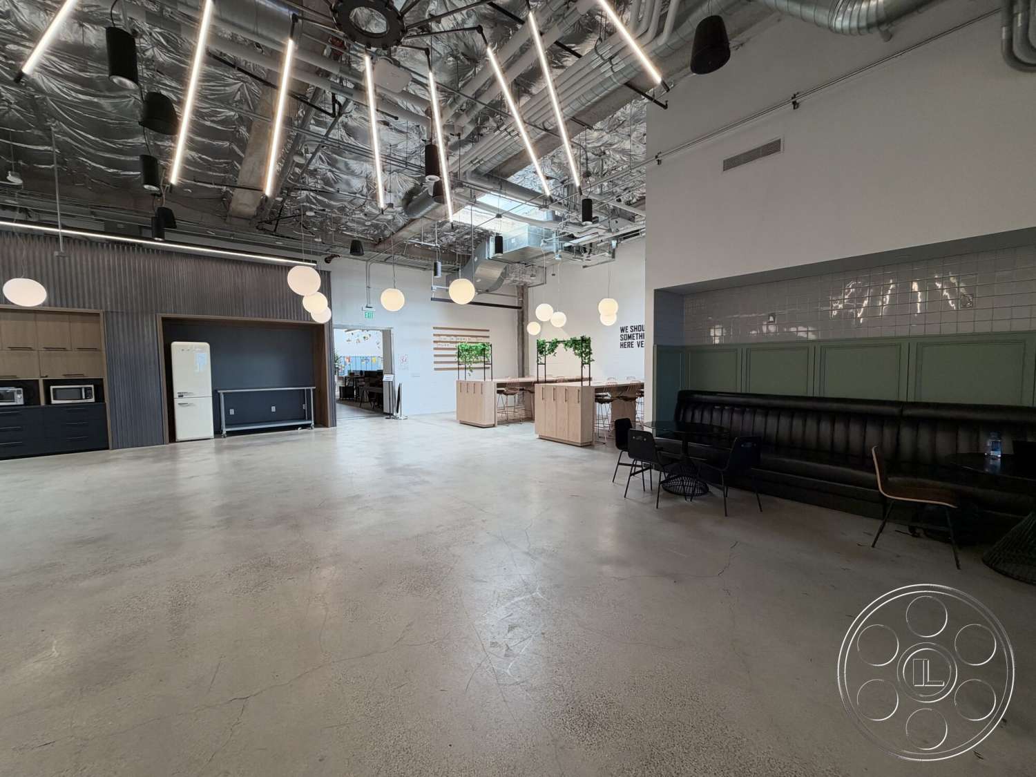 Office 130 - modern pendant lighting, green accent wall, wooden bar counter, open floor plan interior, exposed concrete flooring, industrial ductwork, black kitchen cabinets, metal ceiling panels, white tiled kitchen backsplash, leather banquette seating, brightly lit communal space