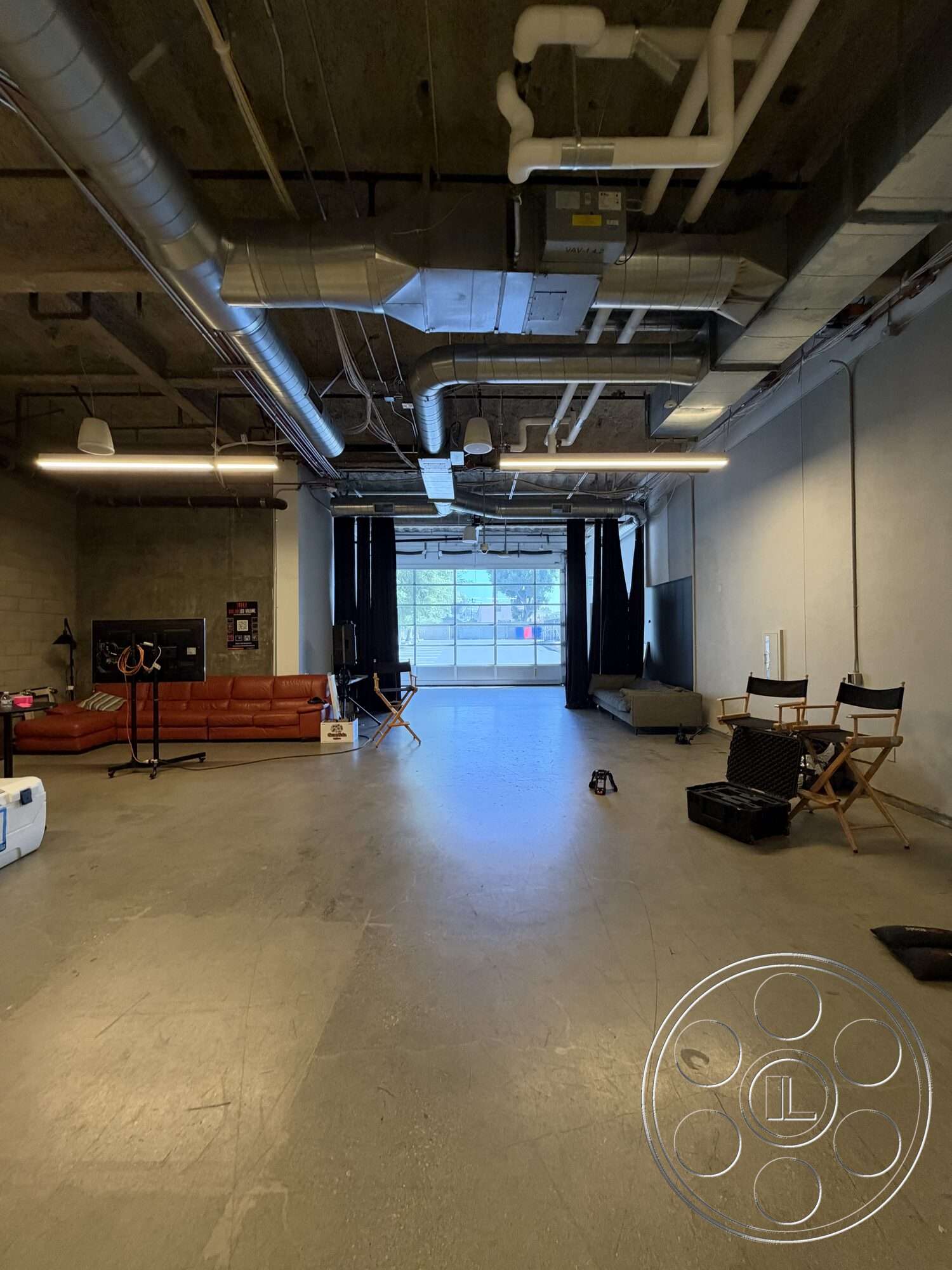 Office 130 - minimalist decor, open floor plan, soft seating, concrete floor, large glass windows, brick wall, industrial lighting fixtures, black curtains, modular seating area, exposed ceiling pipes, folding director chairs