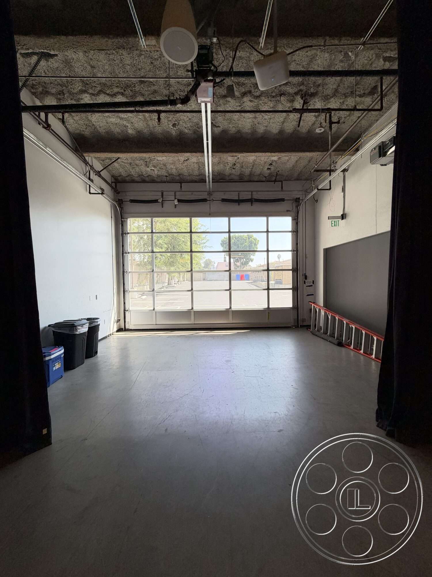 Office 130 - polished concrete flooring, open space layout, exposed concrete walls, large glass garage door, exposed ductwork ceiling, industrial style interior, black curtain partition, ceiling-mounted track lighting, exterior view with greenery
