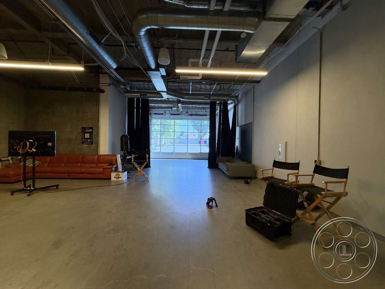 Office 130 - polished concrete flooring, open space layout, exposed concrete walls, large glass garage door, industrial ceiling with exposed ductwork, modern leather sectional sofa, softened lighting details, black curtains for window treatment, wooden director's chairs