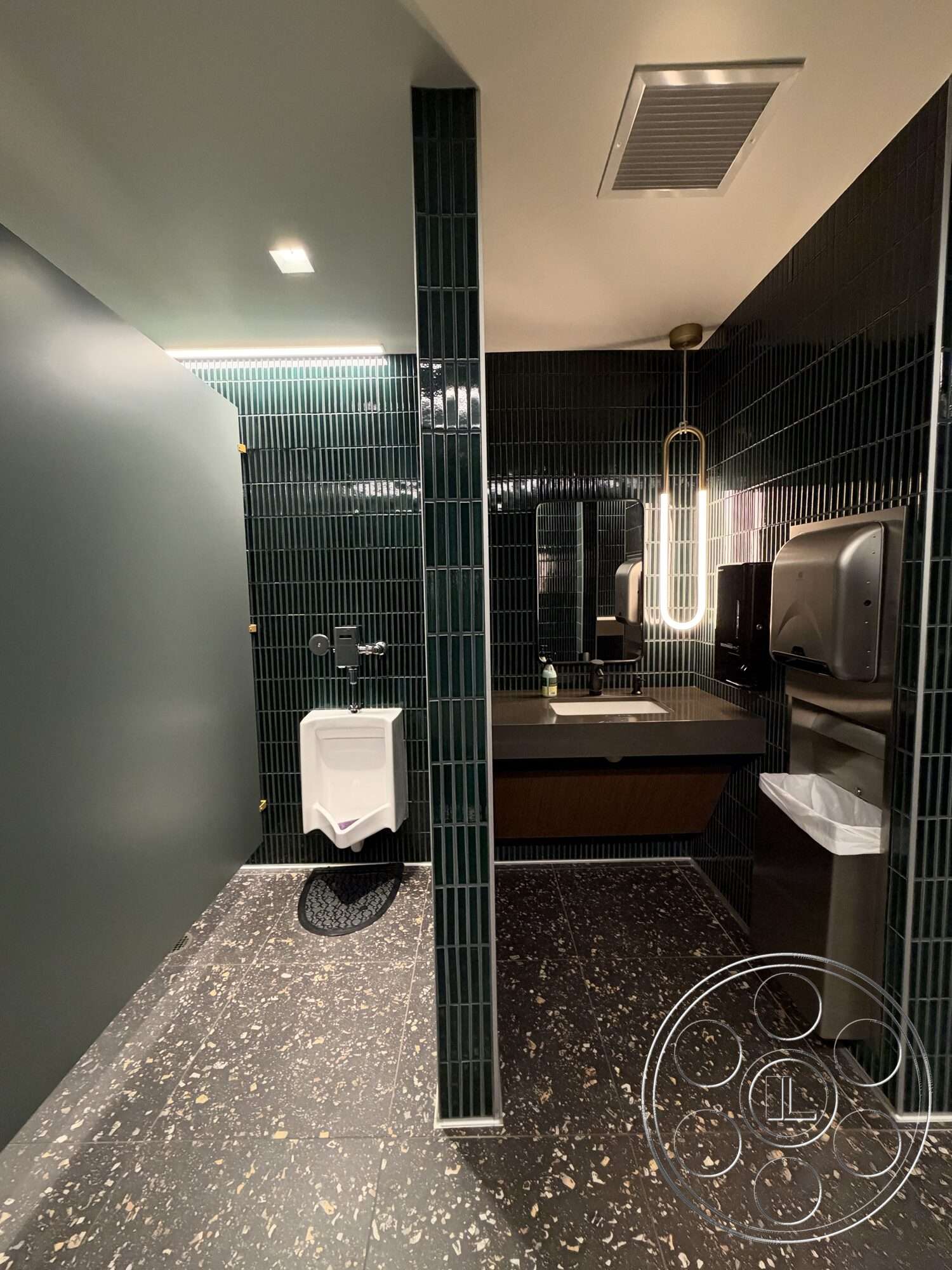 Office 130 - modern light fixtures, textured wall panels, mirrored wall, ambient ceiling lighting, floating bathroom sink, dark green tile bathroom walls, black speckled tile flooring, urinal unit, wooden vanity cabinetry, metal towel dispensers, floor-mounted wastebasket