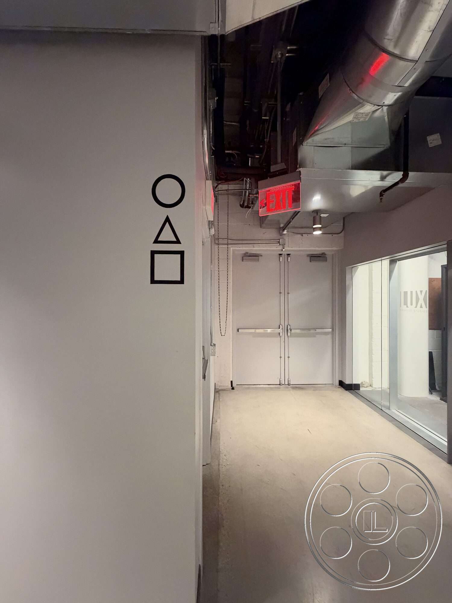 Office 130 - concrete floor, interior hallway, white painted walls, emergency exit sign, metal ductwork ceiling, double glass doors, graphic shapes on wall