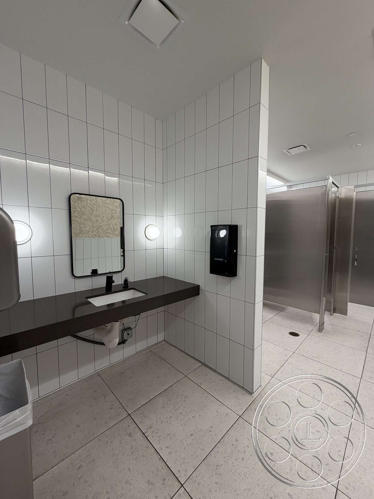 Office 130 - rectangular mirror, sink with faucet, trash can, interior bathroom, stainless steel partition walls, porcelain tile flooring, white ceramic tile walls, modern wall-mounted lighting, dark granite countertop, dispenser for soap