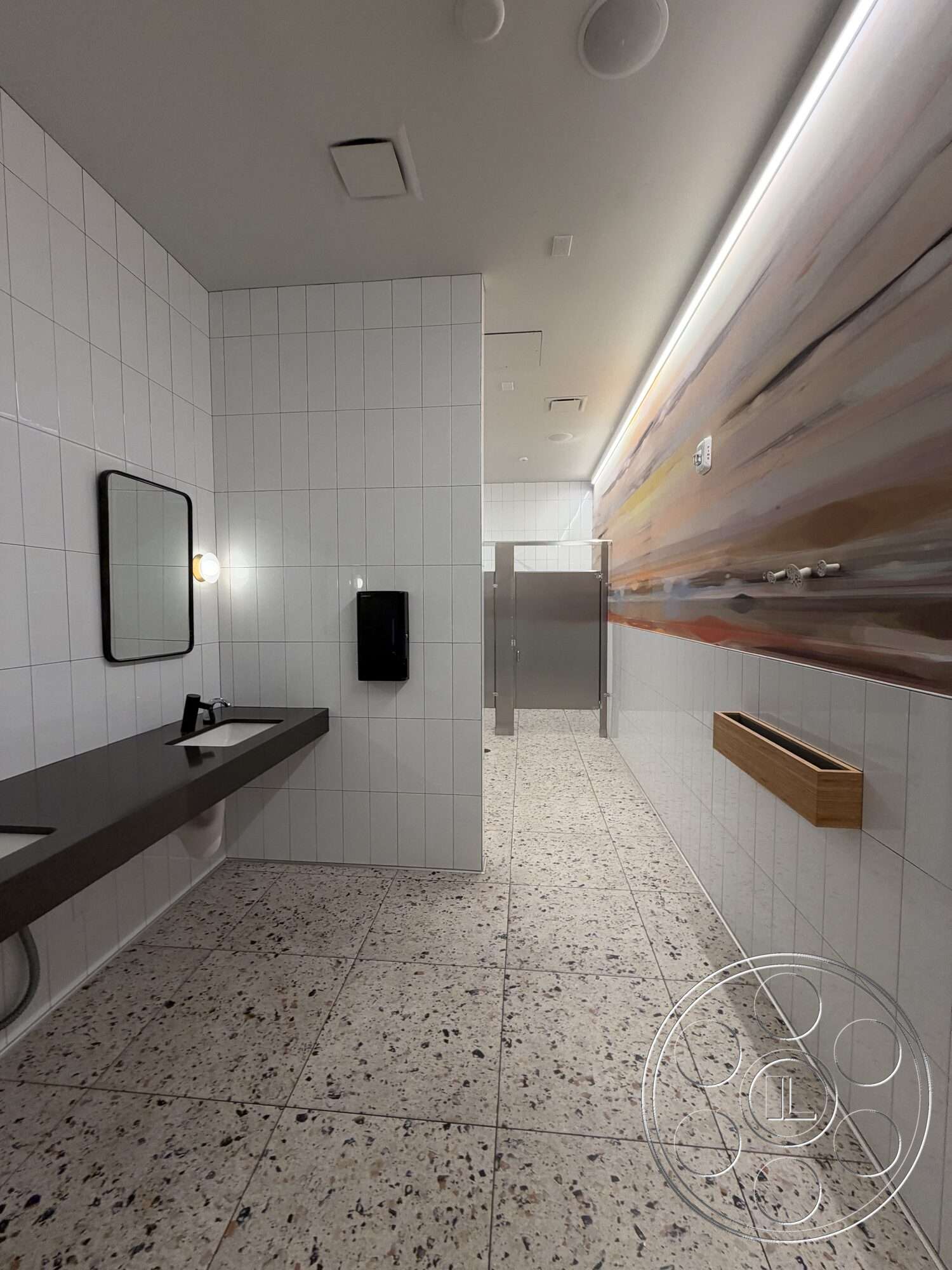 Office 130 - minimalist design, terrazzo flooring, concrete sink countertop, LED recessed lighting, white tile bathroom walls, black wall-mounted soap dispenser, rectangle mirror, built-in wooden shelf, metal bathroom stall doors