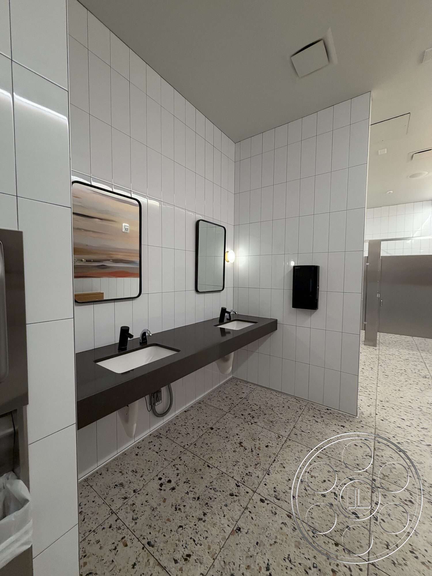Office 130 - minimalist design, terrazzo flooring, modern light fixtures, rectangular mirrors, white tiled walls, interior restroom, black wall-mounted faucets, black granite vanity countertop, double sink fixtures, partitioned stalls