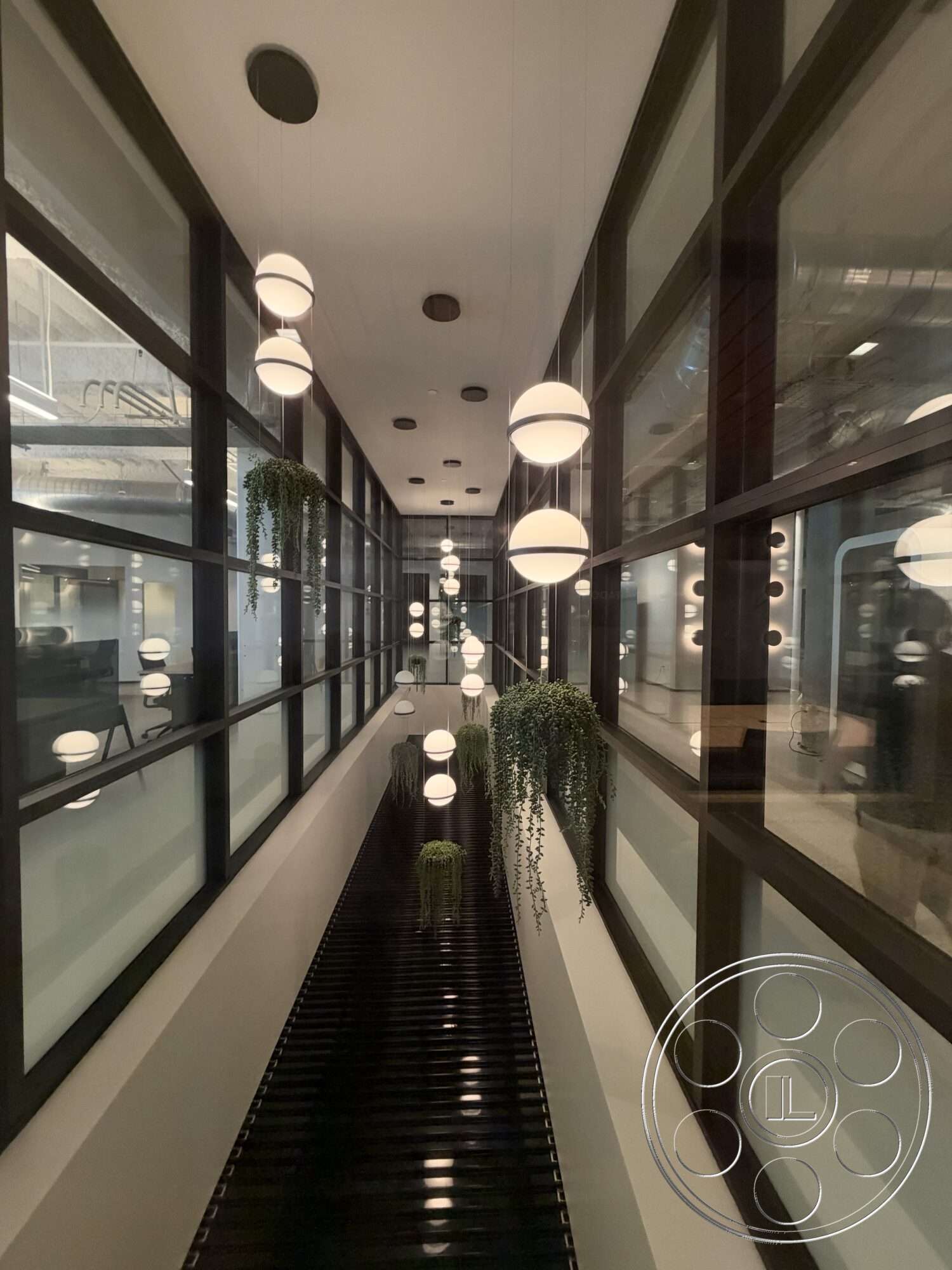 Office 130 - minimalist design, white ceiling, hanging pendant lights, glass partition walls, interior corridor, steel structural elements, rectangular hallway, modern ceiling lights, black tiled flooring, planters with hanging greenery