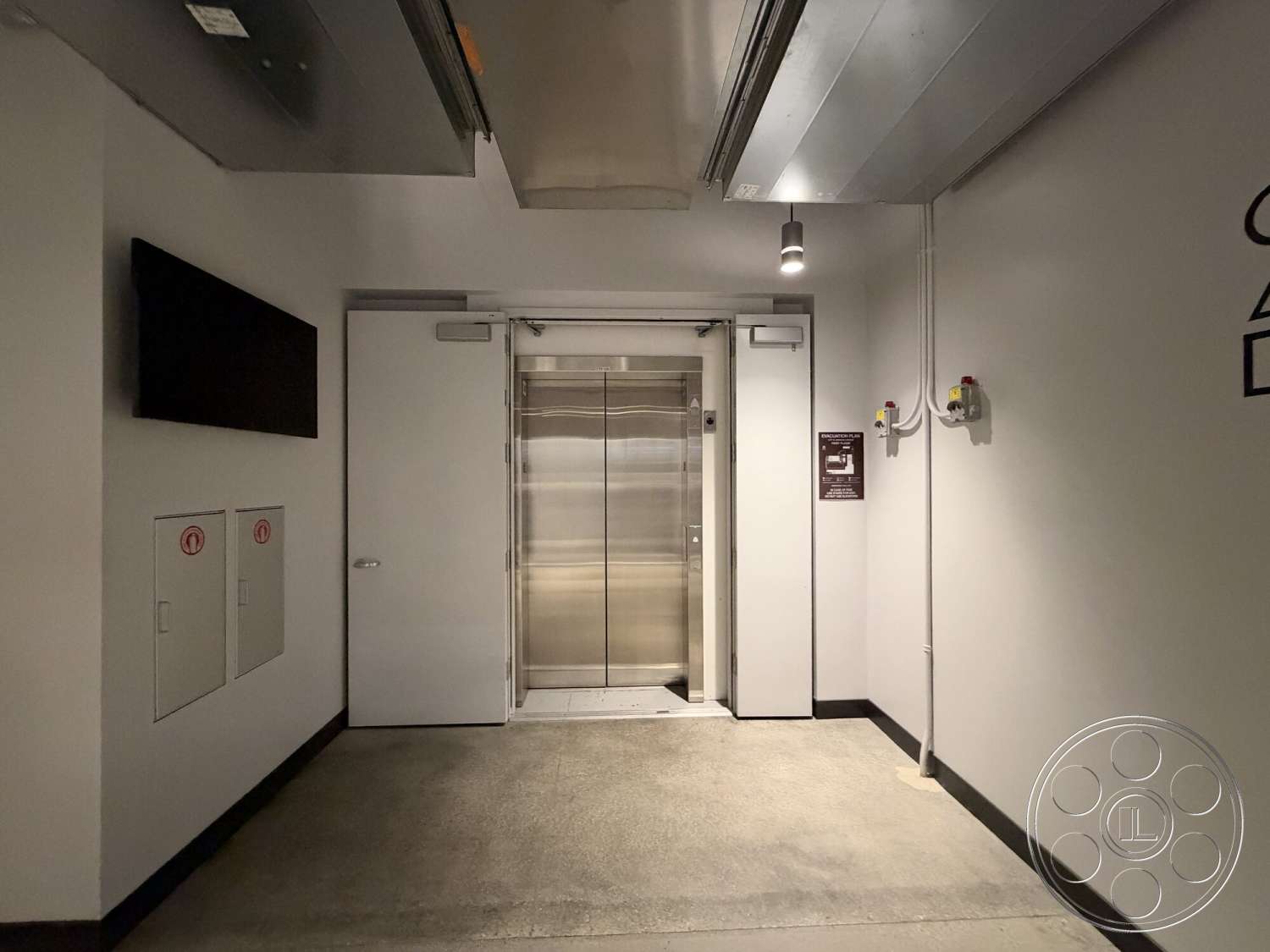 Office 130 - minimalist design, wall-mounted television, concrete floor, flat ceiling, industrial aesthetic, white painted walls, metal elevator doors, exposed ceiling ductwork, electrical control panels, modern hallway