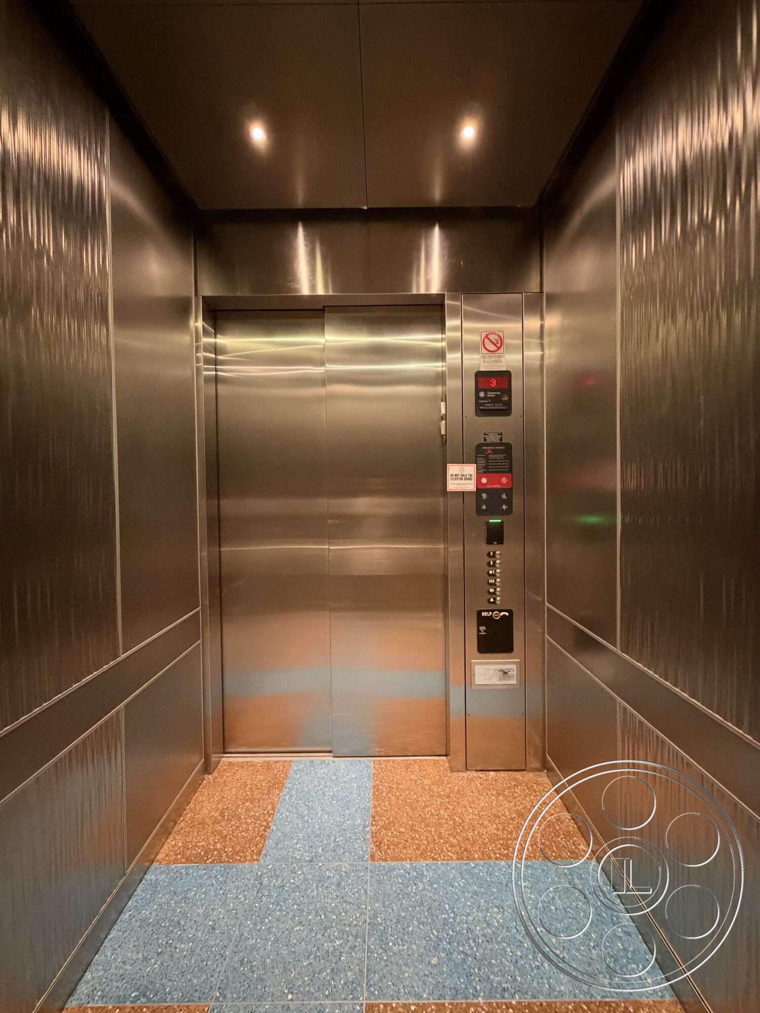 Office 130 - dim ambient lighting, stainless steel walls, interior elevator space, minimalist control panel, glossy stainless steel ceiling, textured paneling, blue terrazzo flooring, metallic elevator doors