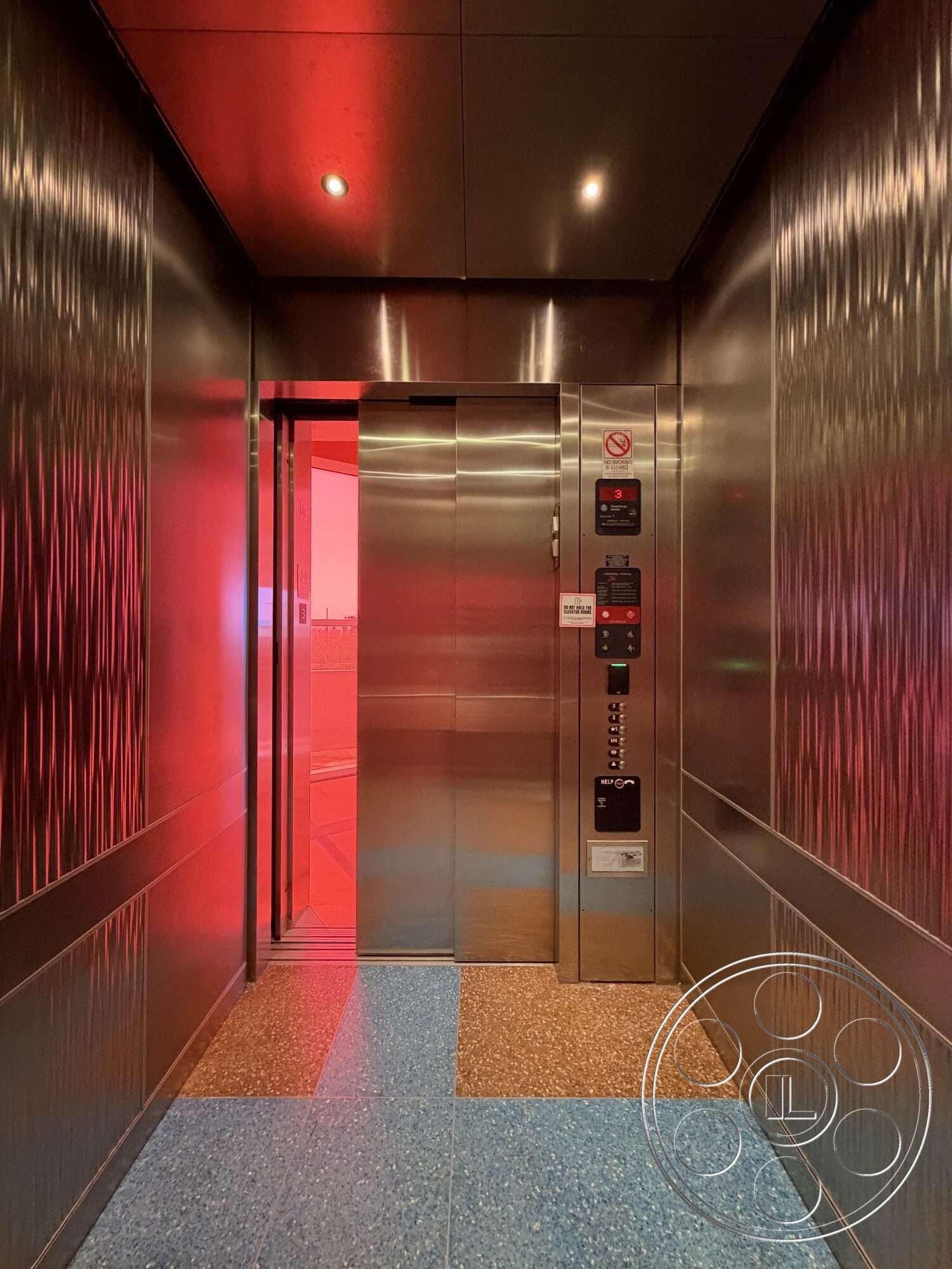 Office 130 - terrazzo flooring, stainless steel elevator doors, elevator control panel, metal wall panels, interior elevator, textured metal wall treatment, glossy blue floor tiles, ambient red lighting, ceiling-mounted lights