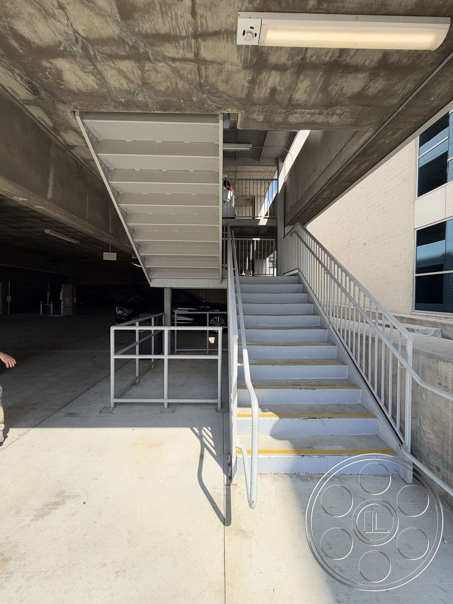 Office 130 - exterior, exposed concrete ceiling, open parking area, metal stair railings, metal stair treads, concrete parking garage floor, white painted brick wall, straight staircase, security lighting fixtures