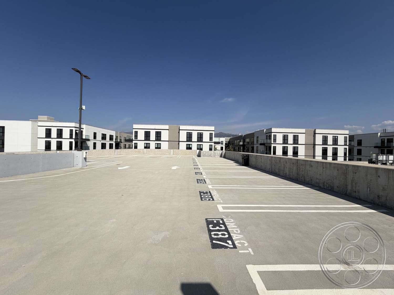 Office 130 - exterior, concrete walls, urban setting, black framed windows, rectangular layout, flat roof design, blue sky backdrop, white painted building facade, exposed concrete parking garage floor, modern apartment building exterior, wide open parking area, street lighting fixtures