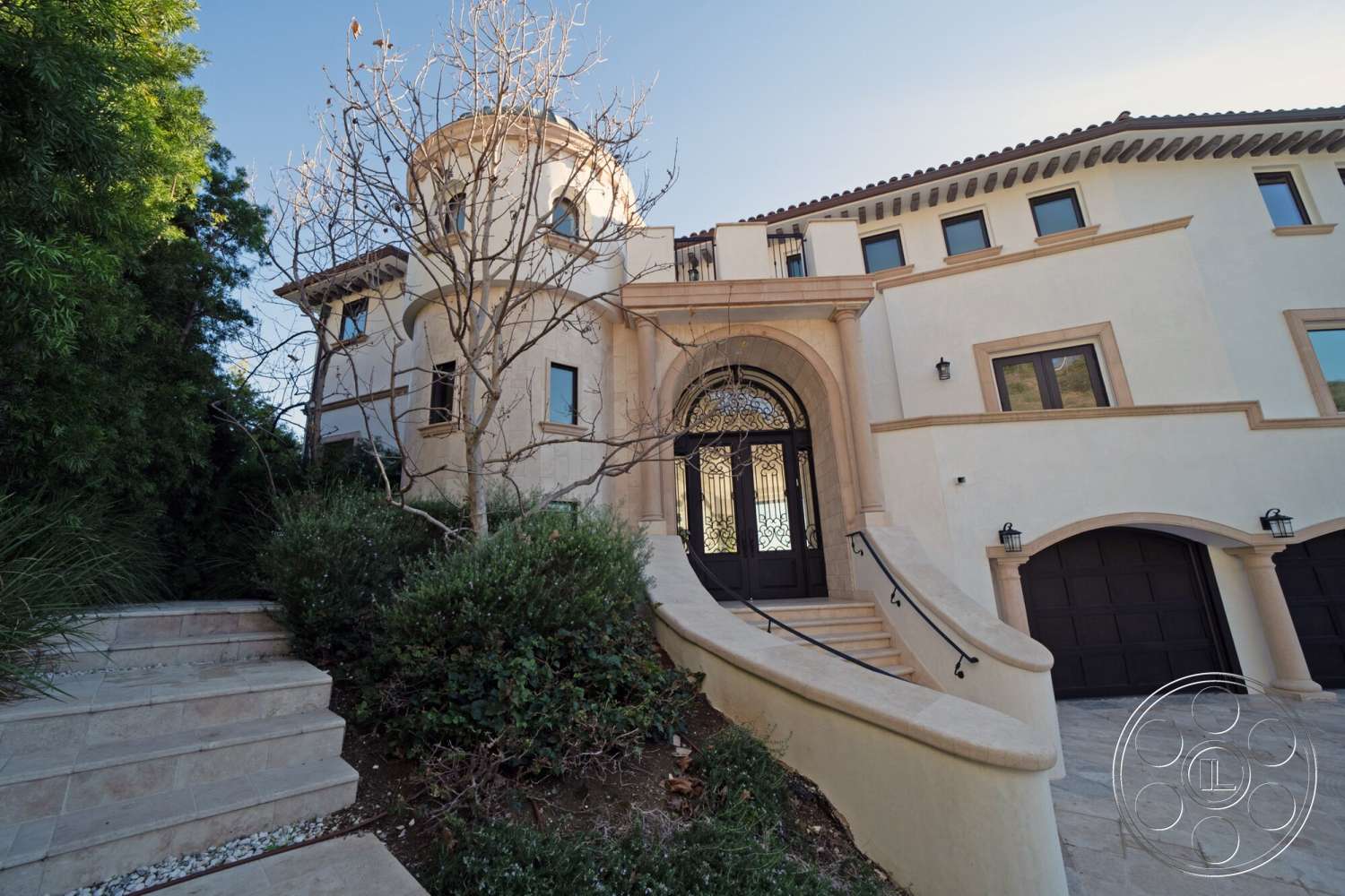 Medit 169 - exterior, circular driveway, gabled roof, stucco walls, arched doorways, stone accents, tile roof, double front doors, slate stone steps, landscaped garden beds, decorative wrought iron details, circular entrance tower, whitewashed exterior