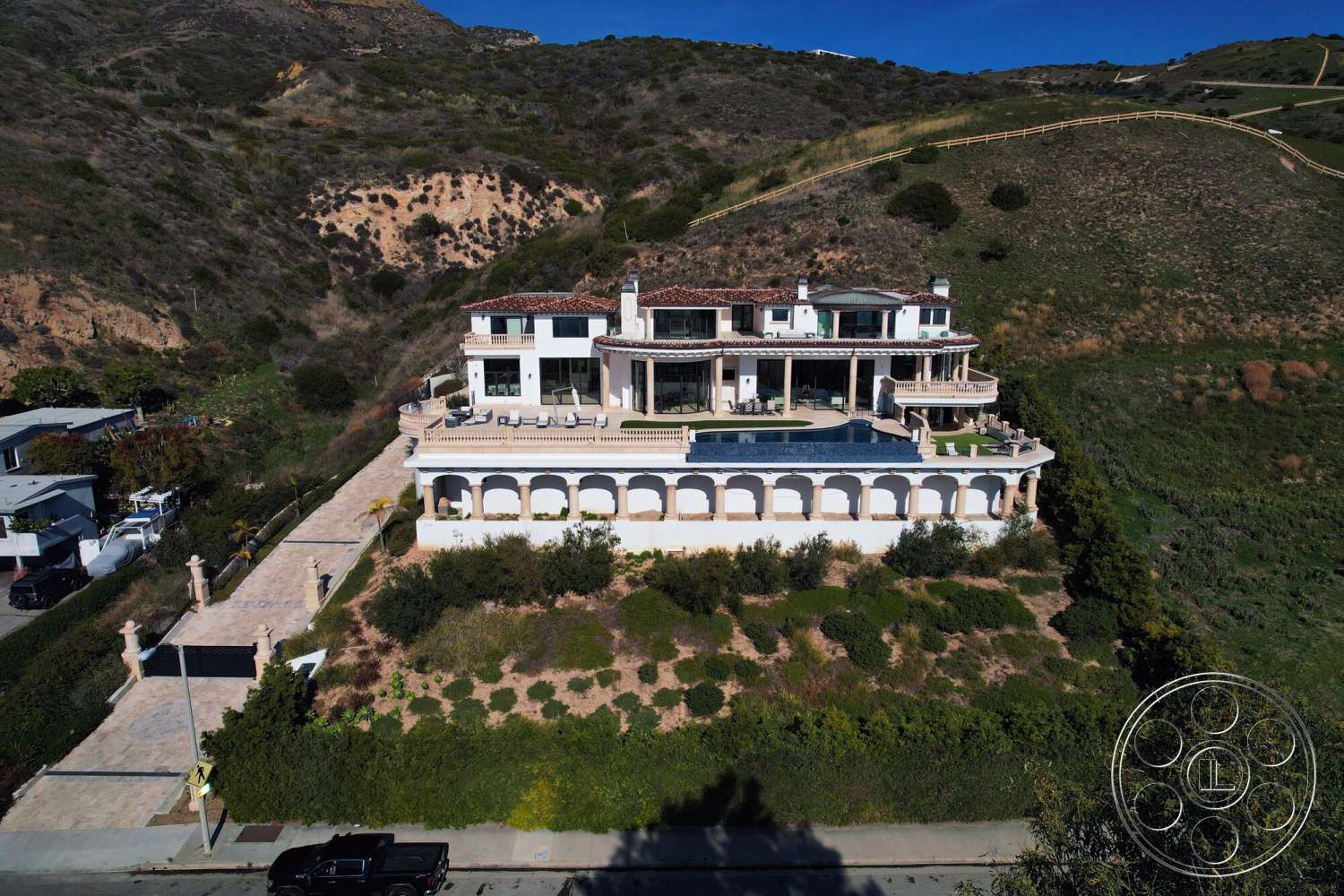 Medit 169 - exterior, paved driveway, white stucco walls, rectangular swimming pool, floor-to-ceiling windows, ocean view, terracotta roof tiles, arched doorways, two-story home, modern Mediterranean style, stone patio flooring, landscaped hillside environment, balustrade railings, curved balcony, rounded columns, expansive outdoor terrace