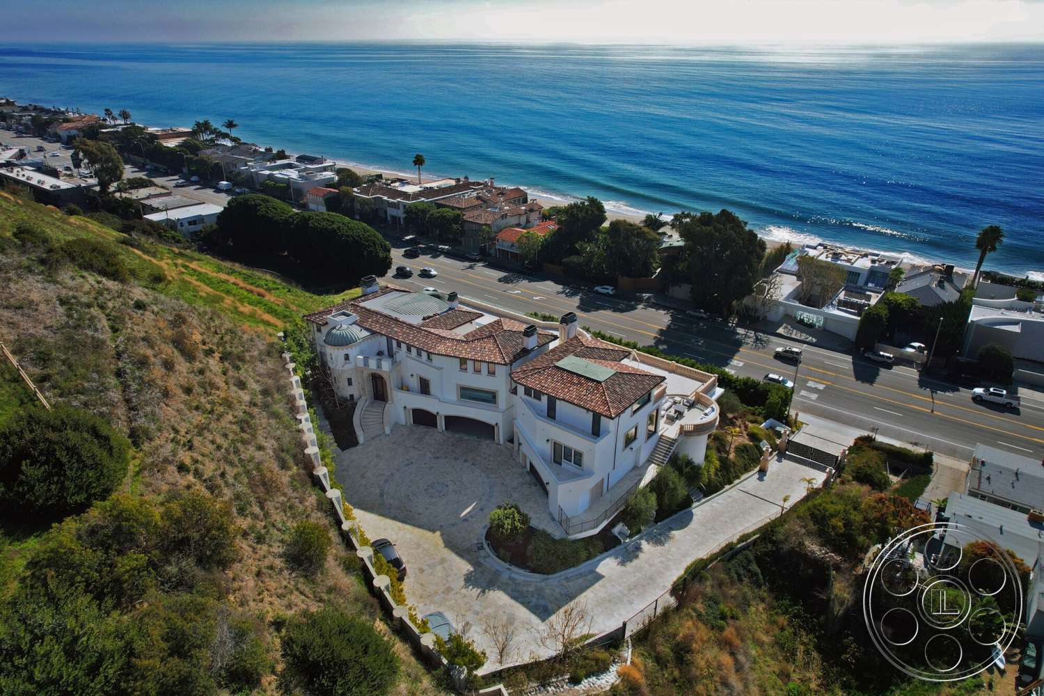 Medit 169 - exterior, landscaped garden, floor-to-ceiling windows, circular driveway, paved walkway, mountain backdrop, stucco walls, arched entryway, red clay tile roof, terraced landscaping, stone patio flooring, glass balcony railings, coastal oceanfront setting, multiple gabled rooftops
