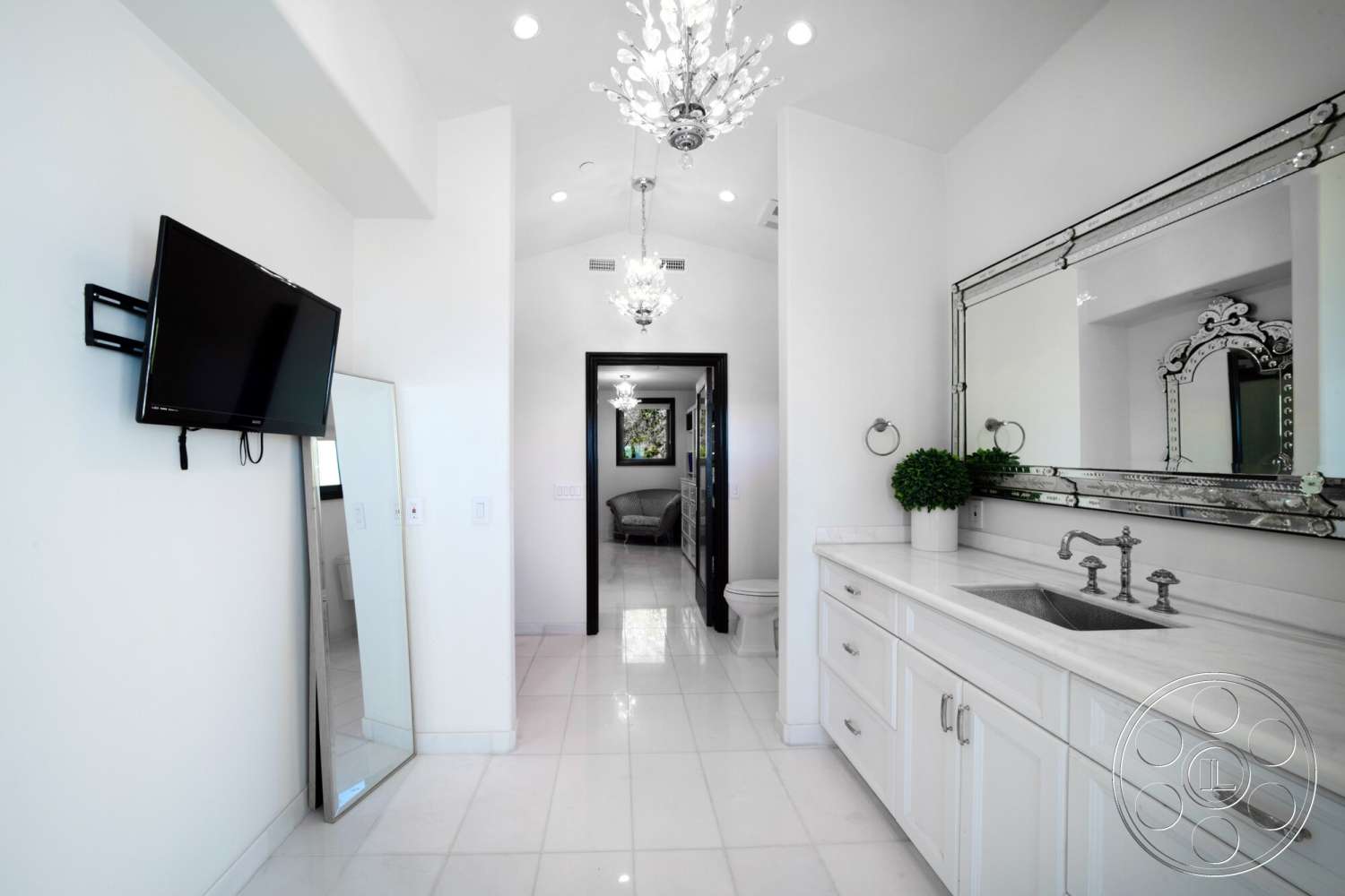 Medit 169 - plant decor, neutral wall color, chandelier light fixture, modern chrome faucet, exposed ceiling lights, flat-screen wall-mounted TV, large framed mirror, white shaker-style cabinetry, white tiled bathroom floors, polished marble countertop, black door frame, freestanding mirror