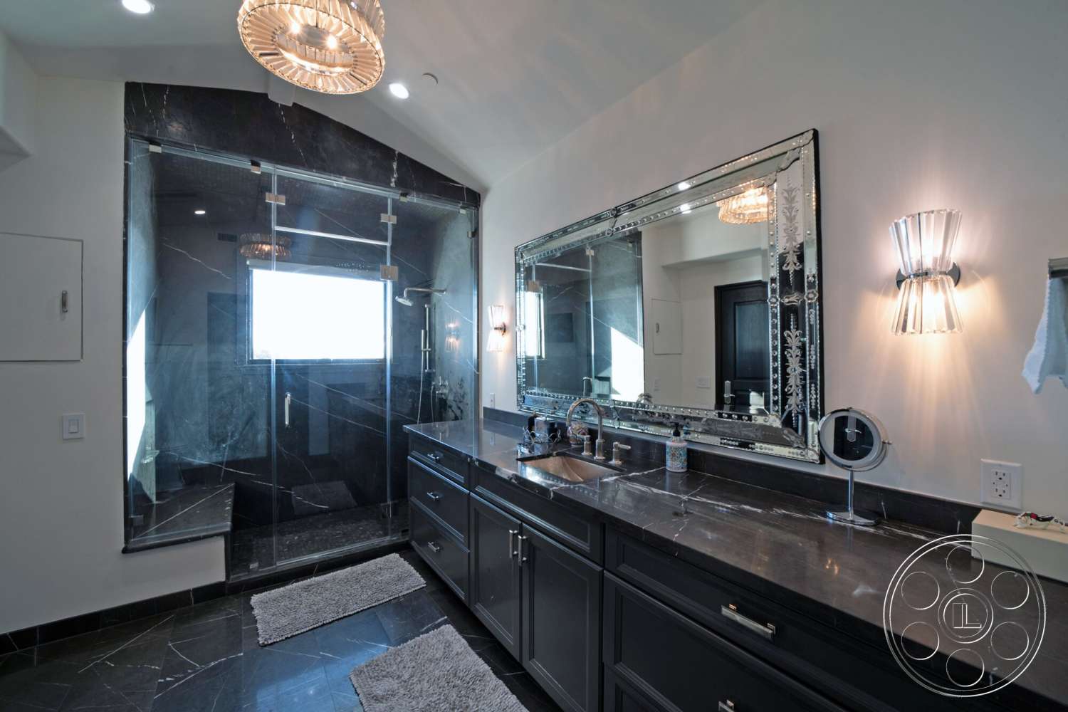Medit 169 - window with natural light, wall-mounted sconces, glass shower doors, chrome faucet fixtures, modern chandelier lighting, luxurious spa-like atmosphere, large decorative mirror, textured white walls, black marble bathroom flooring, black marble shower walls, double sink vanity with black cabinetry, bench seating in shower