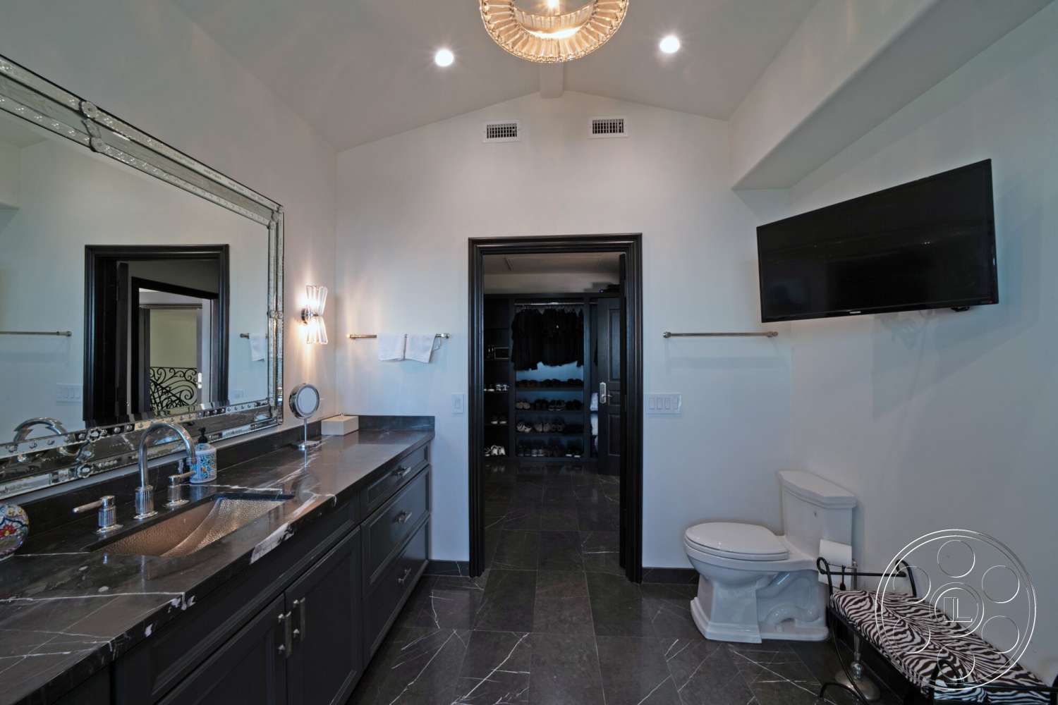 Medit 169 - dark wood cabinetry, wall-mounted TV, mirror with decorative frame, modern bathroom lighting fixtures, white painted walls, white ceramic toilet, black granite bathroom countertops, black door frames, textured slate flooring, double vanity sink, open closet with shelving, zebra-print bench seating