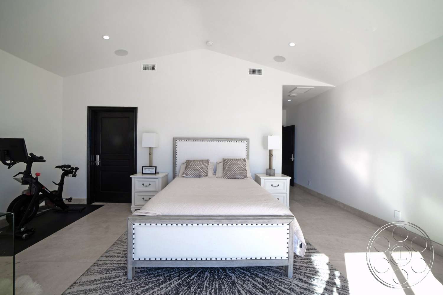Medit 169 - minimalist decor, neutral color palette, textured area rug, light-colored tile flooring, recessed ceiling lighting, white painted walls, black interior door, vaulted bedroom ceiling, modern bedside tables, queen-size bed with upholstered headboard, decorative nailhead trim details, exercise bike on rubber flooring