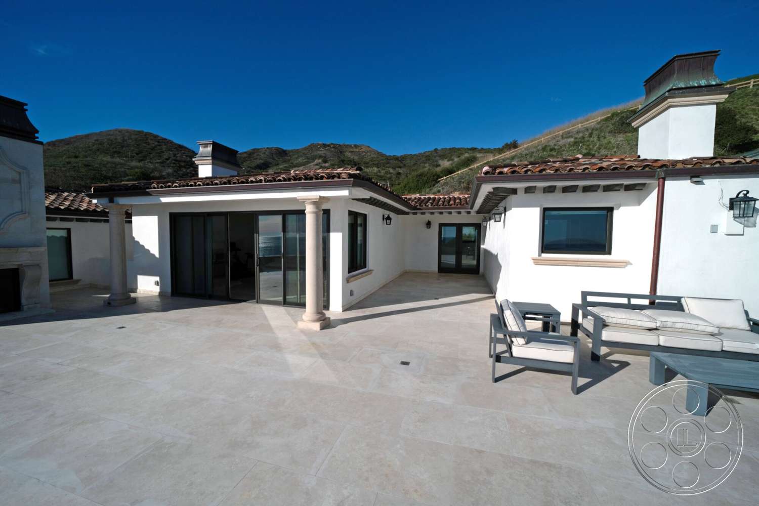 Medit 169 - exterior, white stucco walls, sliding glass doors, terracotta tile roof, modern outdoor furnishings, mountain view setting, stone patio flooring, covered patio area, columns with stone bases