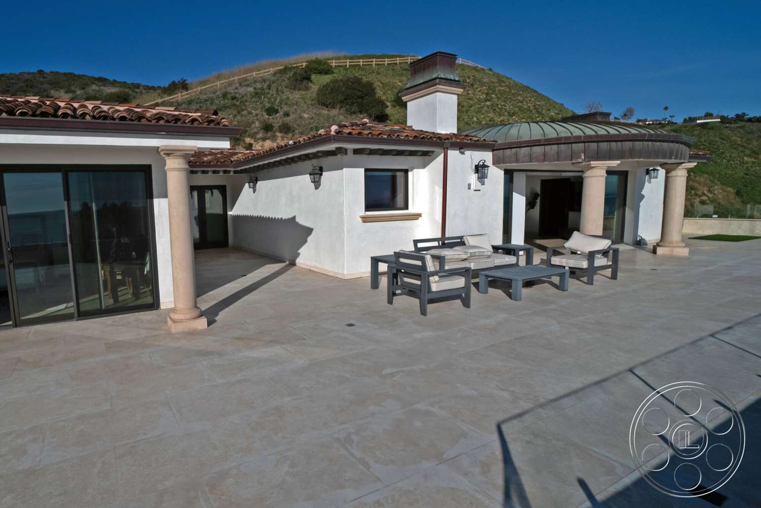 Medit 169 - exterior, glass sliding doors, outdoor seating area, stucco walls, tile roof, concrete patio flooring, columns with stone bases, round arched entrance, landscape hill view