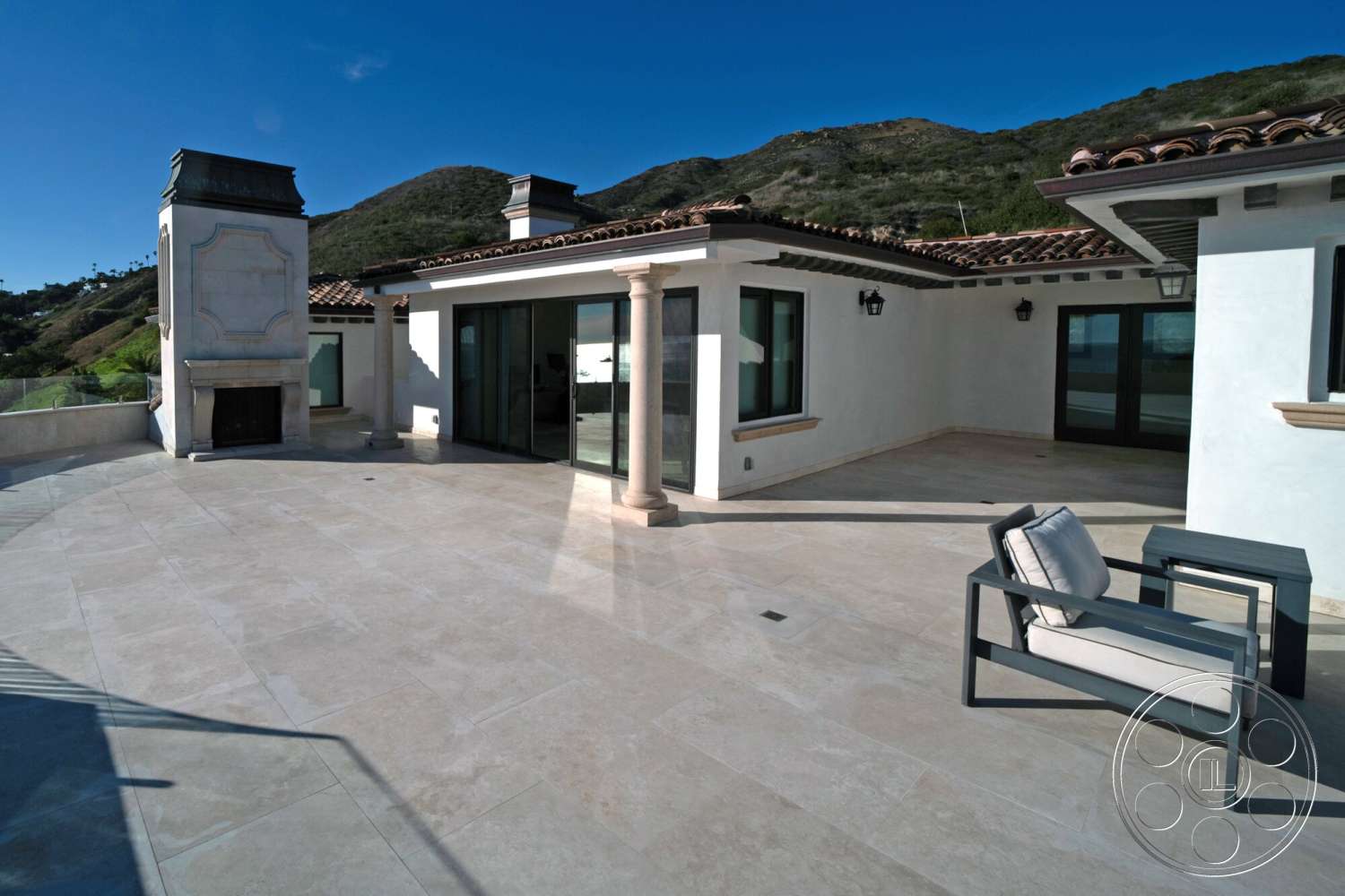 Medit 169 - exterior, large glass sliding doors, terracotta roof tiles, mountain view setting, stucco wall finish, stone fireplace surround, modern outdoor seating area, open patio space, beige stone patio flooring, columned patio structure