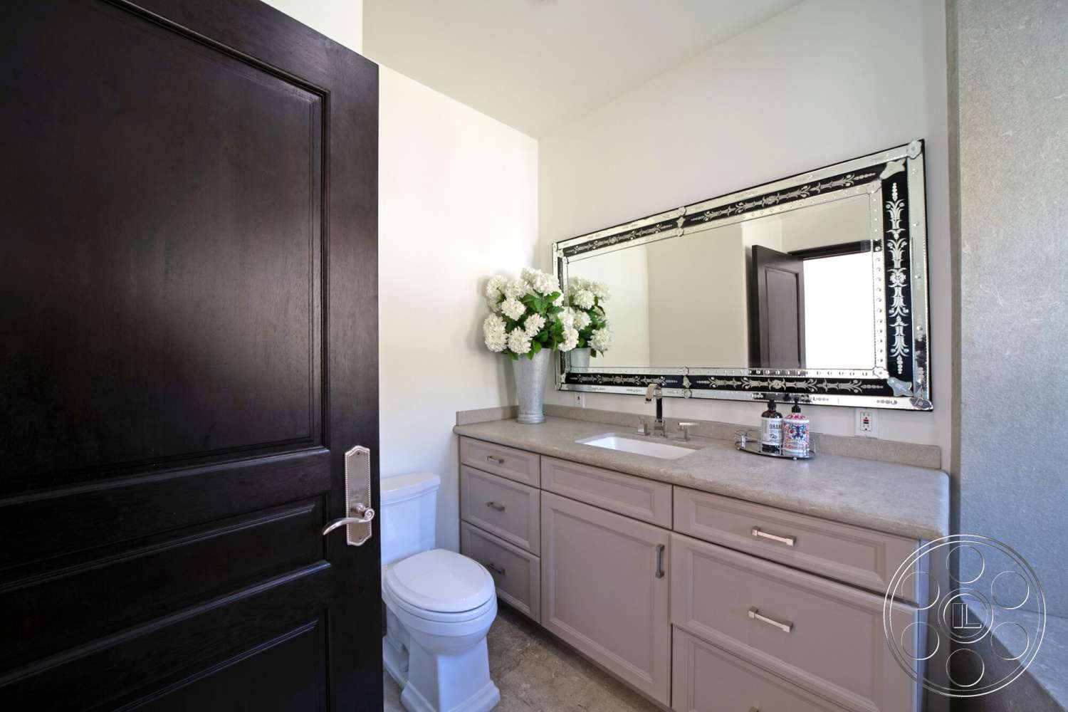 Medit 169 - dark wood door, light-colored painted walls, modern bathroom sink, beige porcelain tile flooring, vanity with light gray countertops, large decorative mirror with black frame, toilet in white ceramic