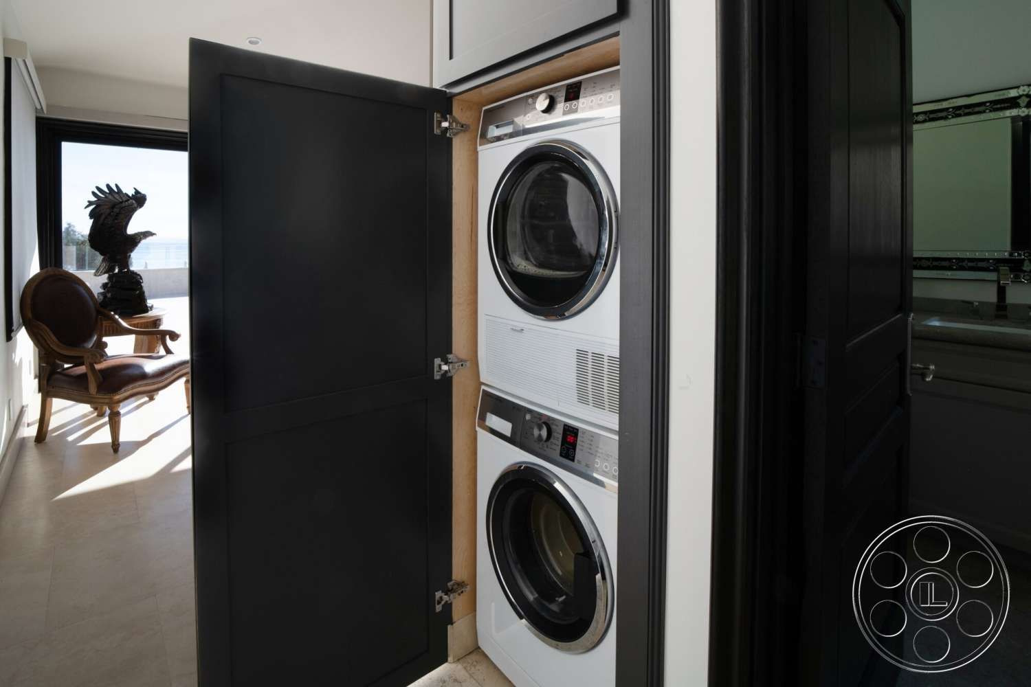 Medit 169 - large windows, natural light, modern design, framed mirror, sculptural decor, light tile flooring, interior laundry room, black cabinet doors, stacked washer dryer units, elegant accent chair