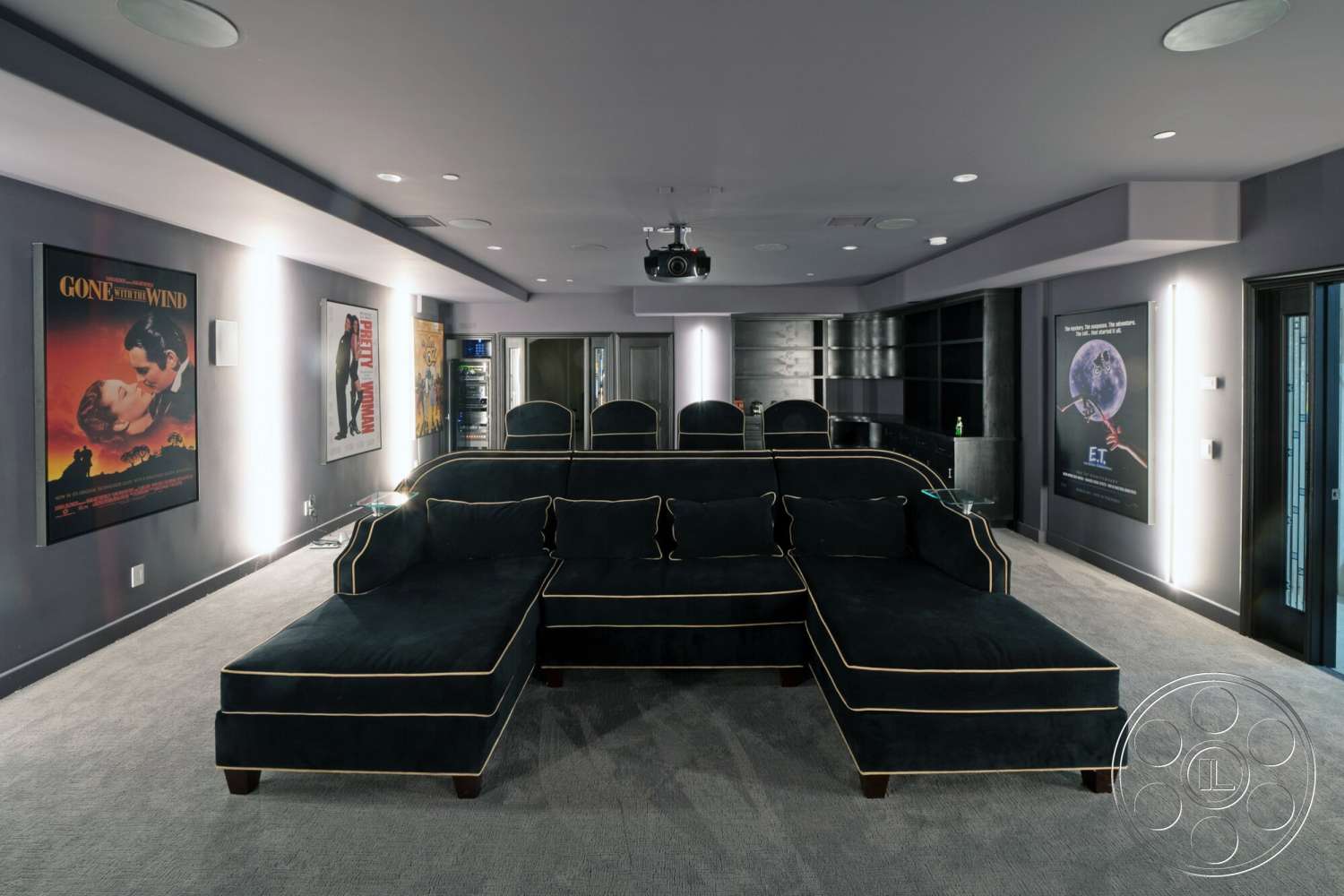 Medit 169 - dark wood cabinetry, ceiling-mounted projector, recessed lighting fixtures, interior cinema room, plush black sectional sofa with gold piping, carpet flooring in cinema room, movie poster wall art, textured gray wall paint, built-in beverage fridge, black accent wall behind seating, spotlight lighting on movie posters, modern home theater design