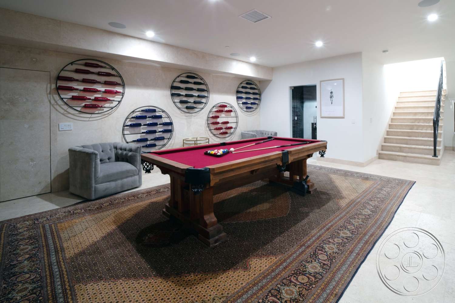 Medit 169 - natural stone flooring, recessed lighting fixtures, neutral painted walls, traditional area rug, metal stair railings, framed artwork on wall, interior game room, tan travertine floor tiles, custom wood pool table, red felt pool table top, gray tufted armchair, decorative wall wine racks, round wine bottle displays, stairs with beige carpet runners, lightly textured wall finishes
