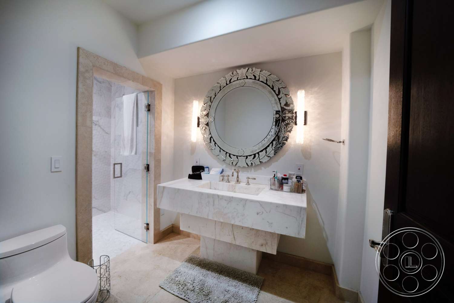 Medit 169 - natural light, textured area rug, glass shower enclosure, stone flooring, modern wall sconces, tile shower walls, framed mirror with decorative detailing, marble bathroom vanity, toilet with a minimalist design, wooden door with dark finish